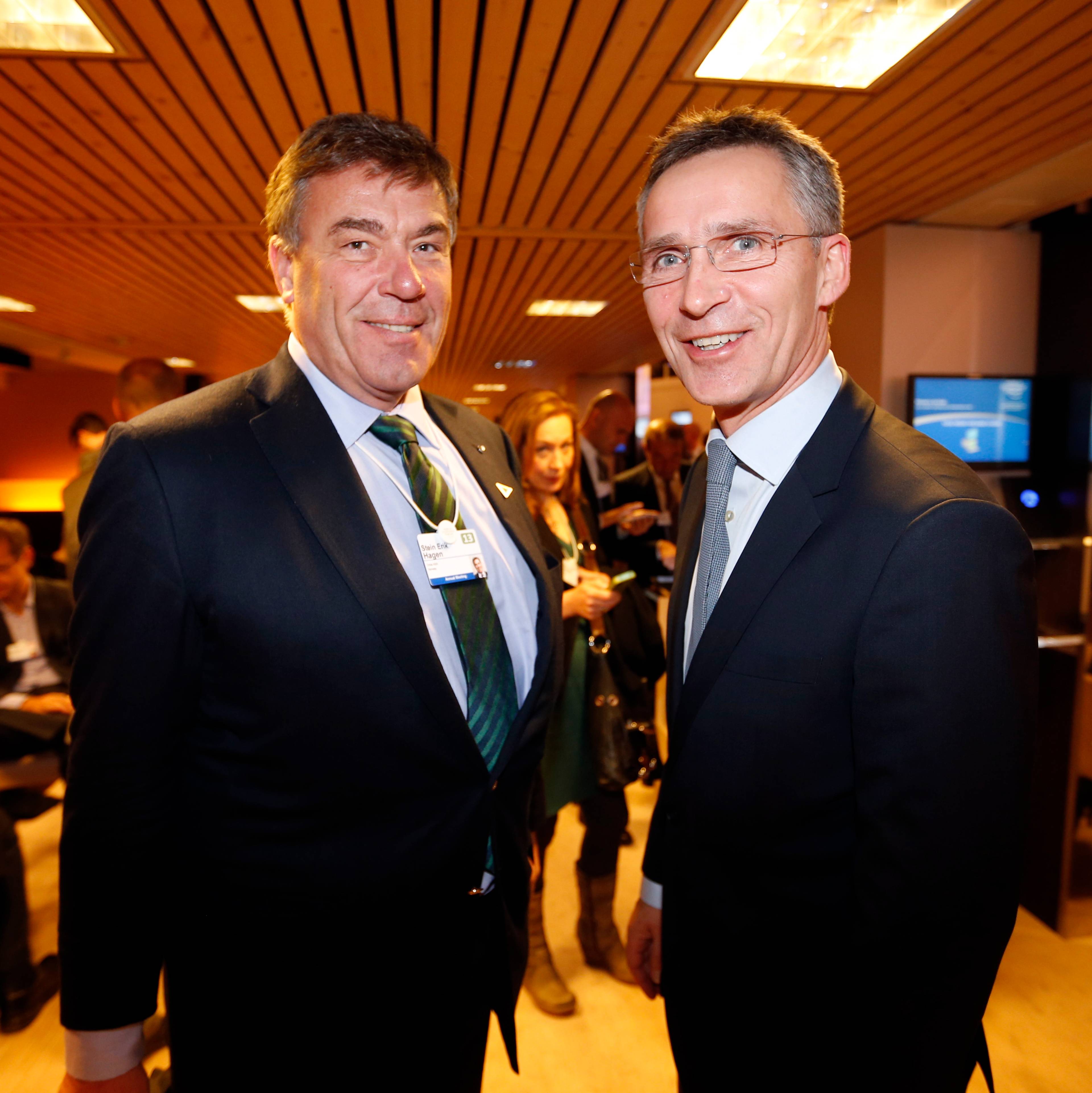 To menn i dress står og smiler foran et publikum. Den ene har en grønn slips, den andre en lyseblå slips. (Bildebeskrivelsen er laget av en KI-tjeneste)