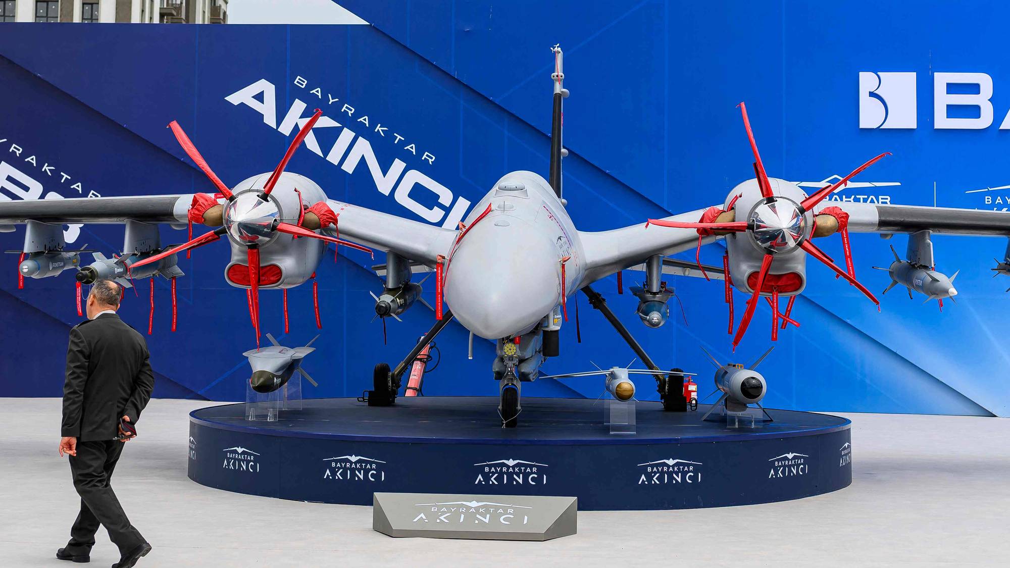 En mann i mørk dress går forbi et utstillingsområde. Der står det en stor, hvitt og grått drone med to propeller. Bak flyet er en stor, blå utstillingsvegg med logoen til Baykar og navnet på flyet, Akinci.