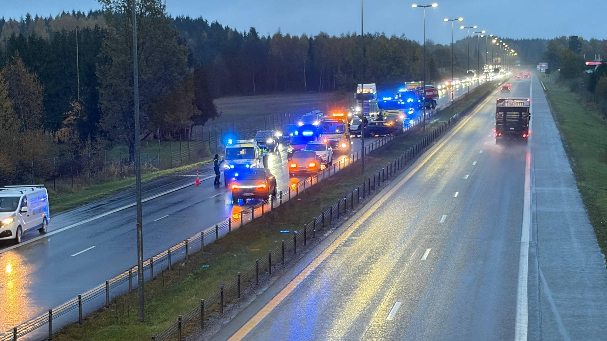 Nødetater på ulykkessted på våt motorvei med kø og blinkende blålys i skumringen. (Bildebeskrivelsen er laget av en KI-tjeneste)