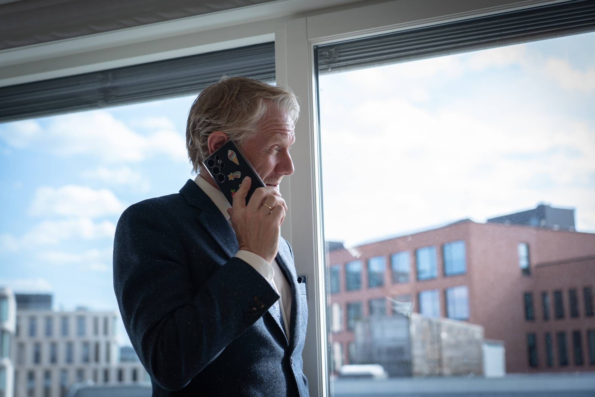 En eldre mann i dress snakker i telefonen ved et vindu. Utenfor ses bygninger og en blå himmel. Telefonen har flere klistremerker. (Bildebeskrivelsen er laget av en KI-tjeneste)