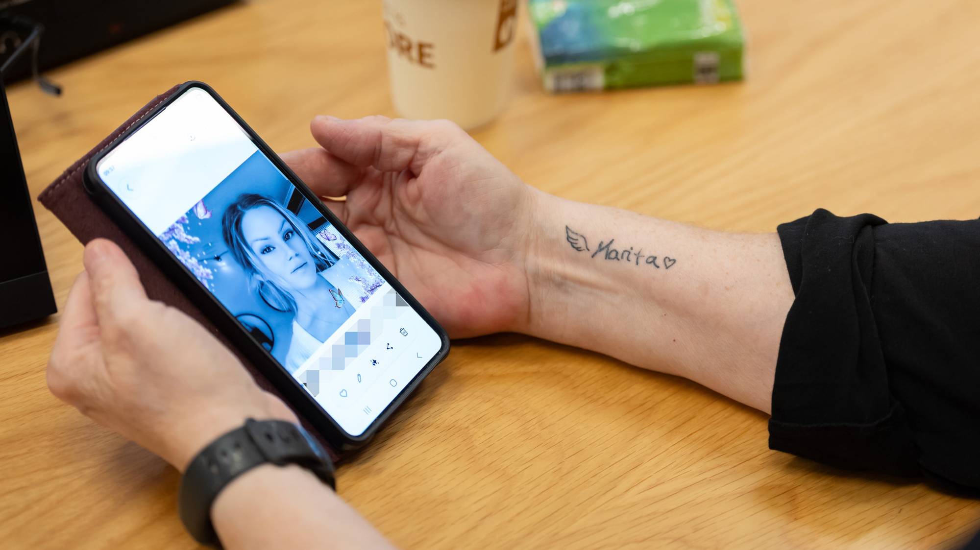 Mor til avdøde Marita Løfshus Haugen sitter i en rettssal i Trøndelag tingrett. Hun kikker på et bilde av sin datter på mobiltelefonen. På armen har hun tatovert inn navnet på sin datter. Vi ser kun armene til mor og antydninger til en svart genser. På bordet foran henne en pappkopp og en pakke med papirlommetørkle