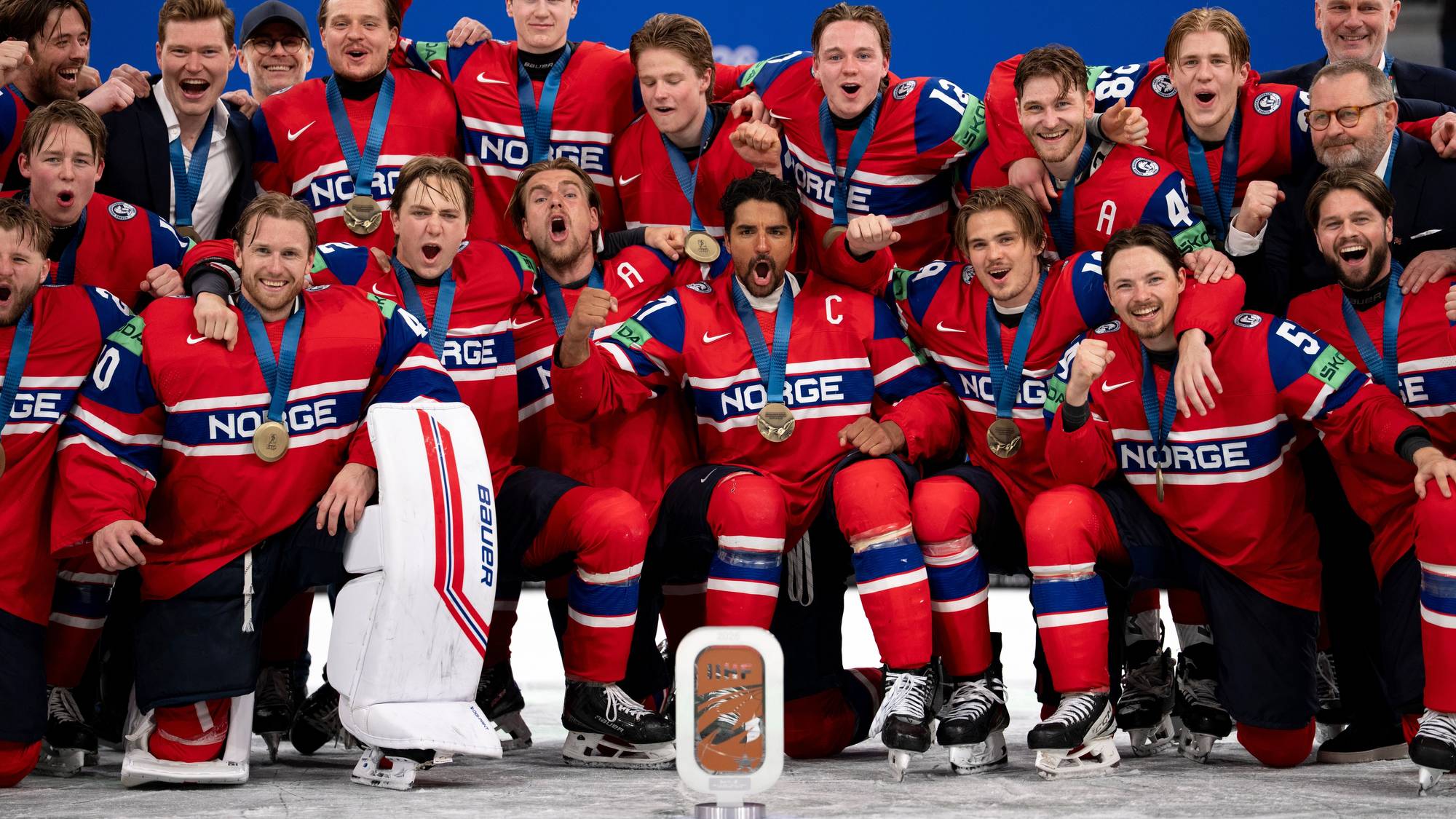 Norges ishockeylag feirer seier med medaljer rundt halsen. Spillerne er kledd i røde drakter med teksten "NORGE". I forgrunnen står en keeper med utstyr. En pokal er plassert foran laget. (Bildebeskrivelsen er laget av en KI-tjeneste)