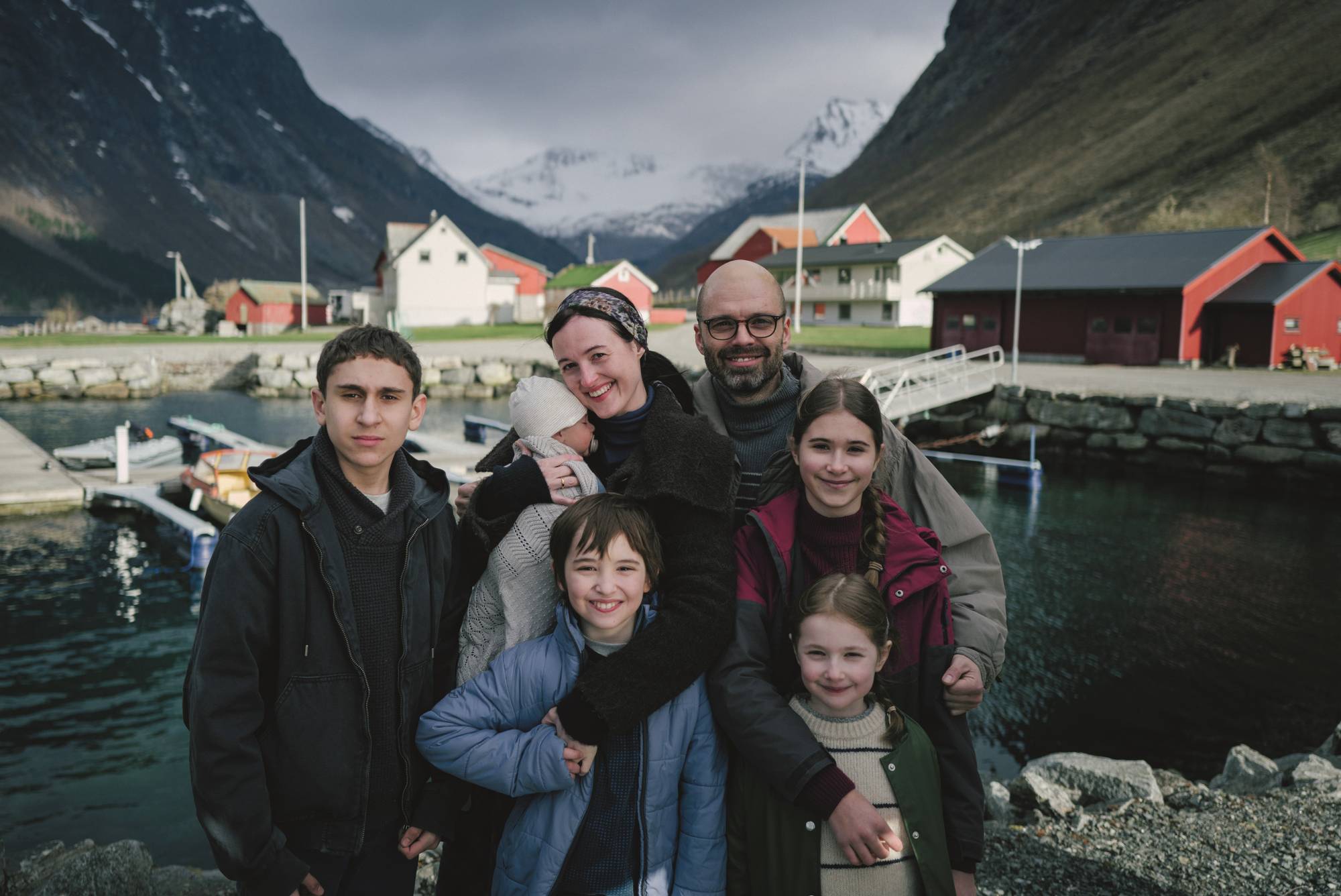 En familie på syv poserer for et bilde ved en havn. En mann og en kvinne står bakerst, med en baby i armene til kvinnen.