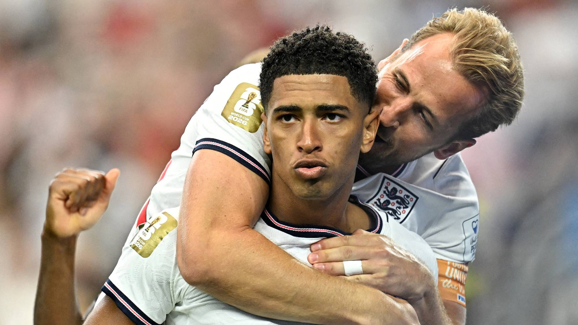 Fotballspillere Jude Bellingham og Harry Kane feirer. Kane gir Bellingham et kyss på kinnet. De er begge iført hvite drakter med Englands flagg på.