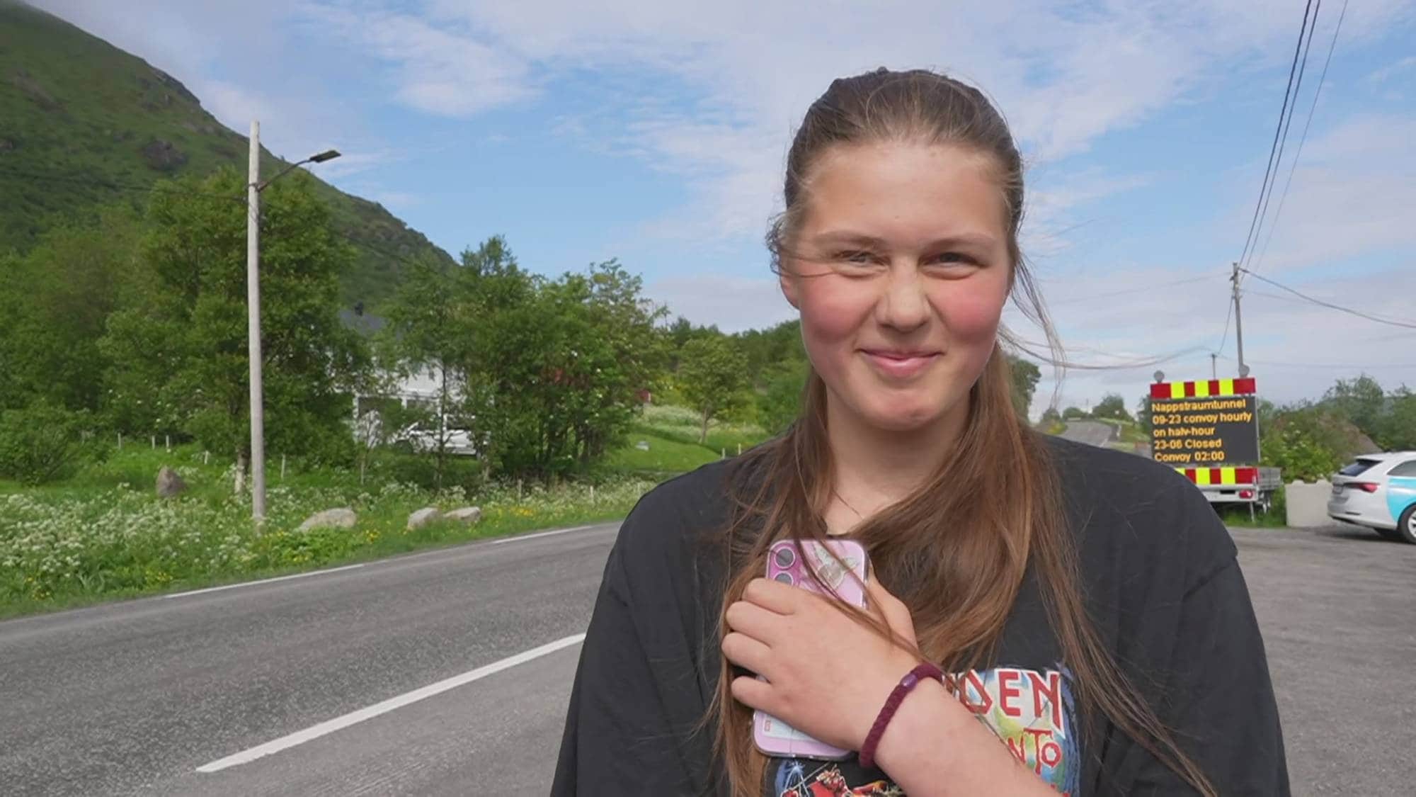 En ung jente holder en mobiltelefon foran seg og smiler. Bak henne er en vei, grønne trær og en fjellside. Til høyre står en skilttruck med informasjon om Nappstraumtunnelen. (Bildebeskrivelsen er laget av en KI-tjeneste)