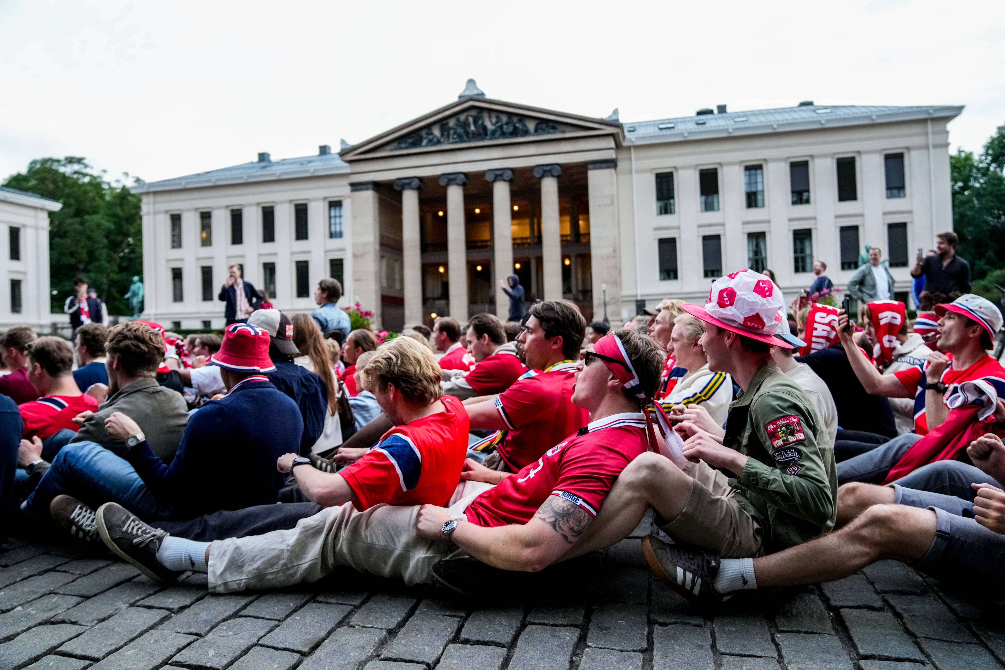 En gruppe mennesker sitter tett på en brosteinsbelagt gate, kledd i rødt og hvitt og med norske flagg. I bakgrunnen ruver en stor, klassisk bygning med søyler. Mange av personene ser ut til å være samlet for å se på en skjerm eller et arrangement. Noen bærer supporterhatter og har malt flagget i ansiktet.