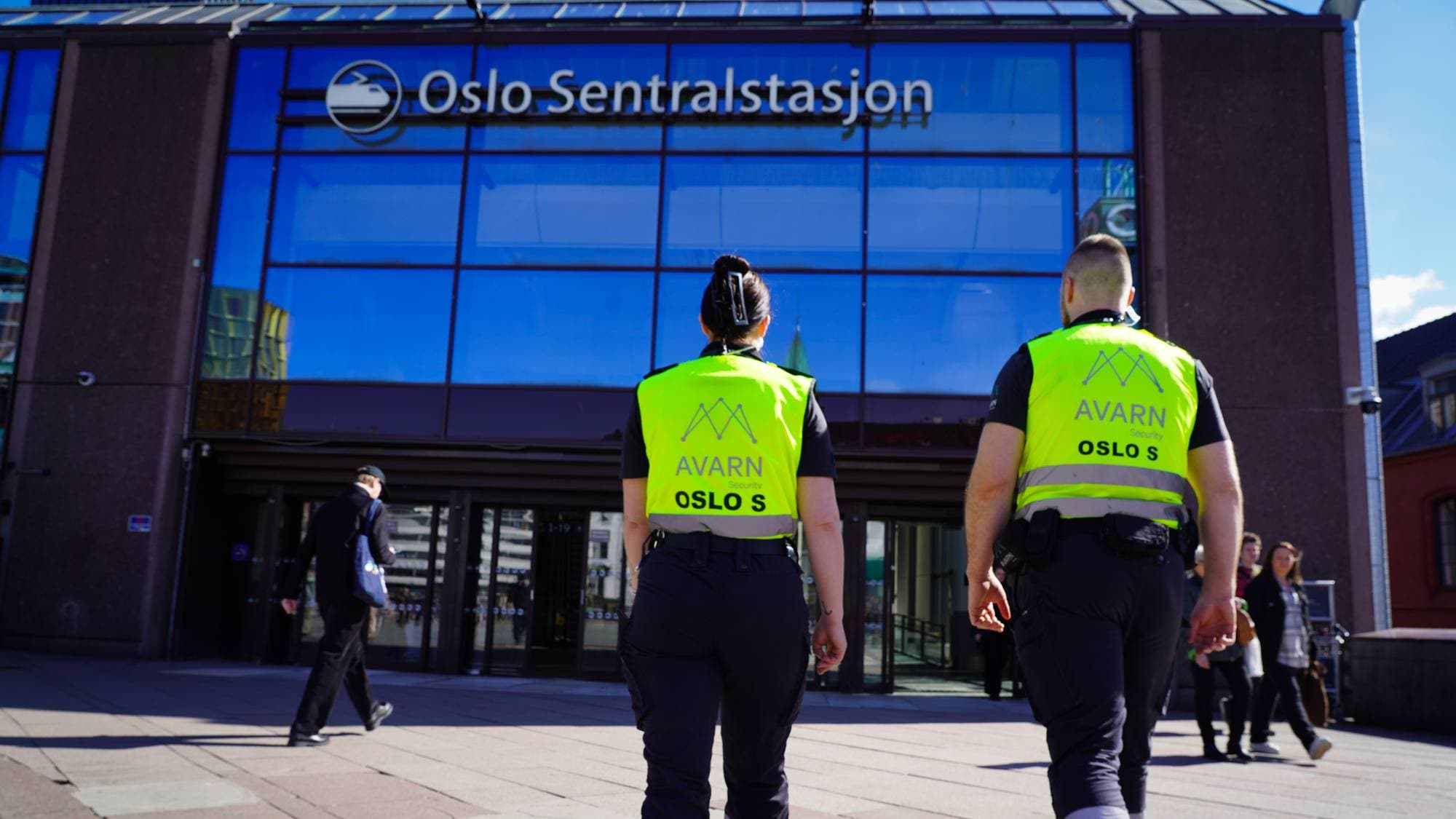 To personer iført gul vest og mørke uniformsbukser går forbi inngangen til Oslo Sentralstasjon. De jobber for AVARN Security. En mann med blå bag går foran dem. (Bildebeskrivelsen er laget av en KI-tjeneste)