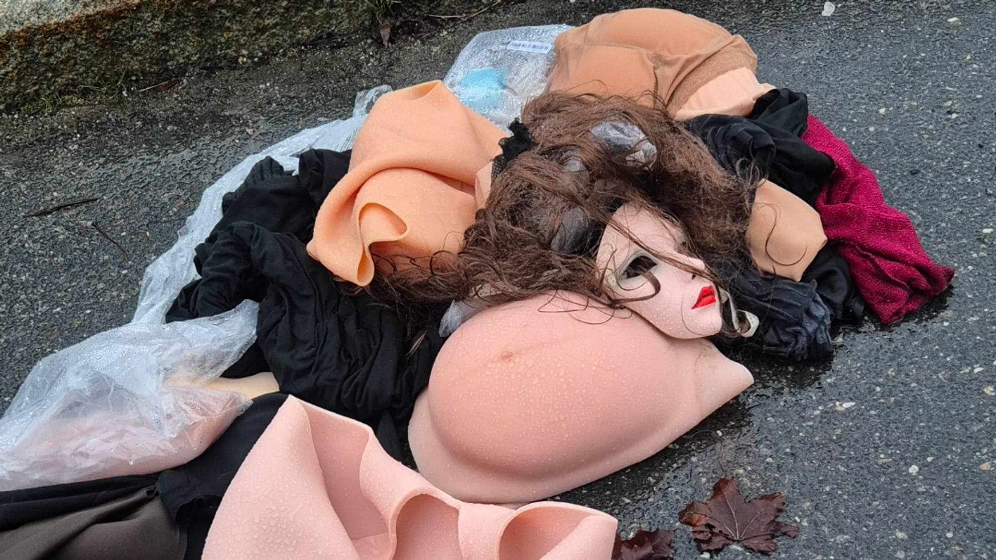 Kastet dukkehode, brystproteser og klær ligger på en våt asfaltholdeplass. (Bildebeskrivelsen er laget av en KI-tjeneste)