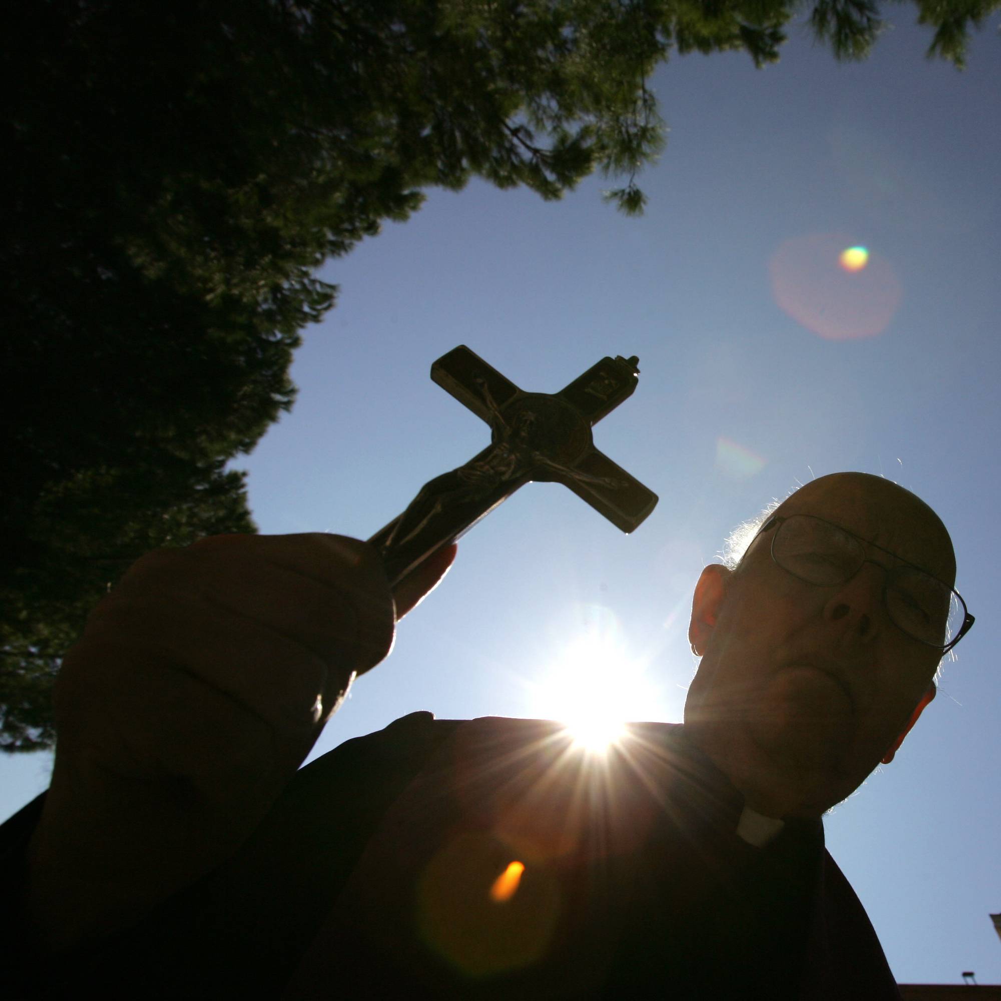 En prest holder opp et krusifiks mot solen.
Presten har på seg prestekjole og briller.
Det er trær på sidene og en bygning i bakgrunnen.
Solen skinner sterkt på himmelen. (Bildebeskrivelsen er laget av en KI-tjeneste)