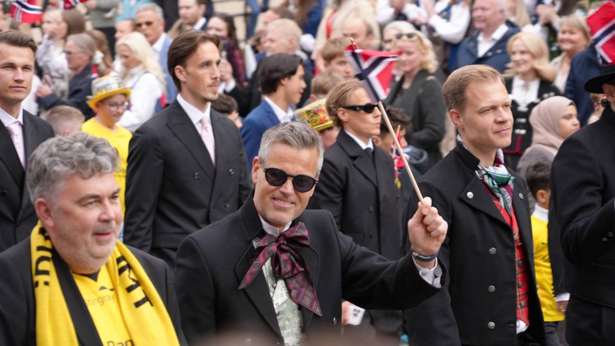 En gruppe menn i dress og bunad deltar på en begivenhet. En av mennene holder et lite norsk flagg. Han har på seg solbriller og smiler til kameraet. Det er mange tilskuere i bakgrunnen. (Bildebeskrivelsen er laget av en KI-tjeneste)