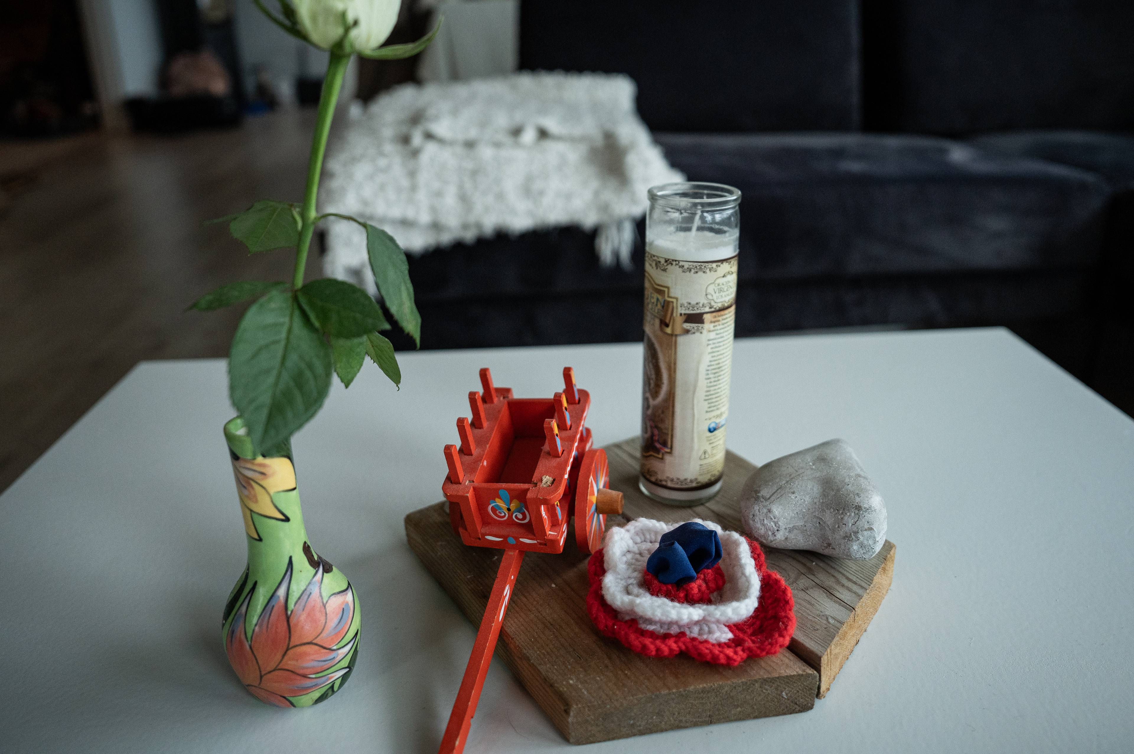Et hvitt rosestilk i en dekorert vase står på et hvitt bord. Ved siden av står et lite rødt trevogntog, en glasslykt, en stein og en rød og hvit heklet blomst. I bakgrunnen sees en sofa og et hvitt pledd. (Bildebeskrivelsen er laget av en KI-tjeneste)