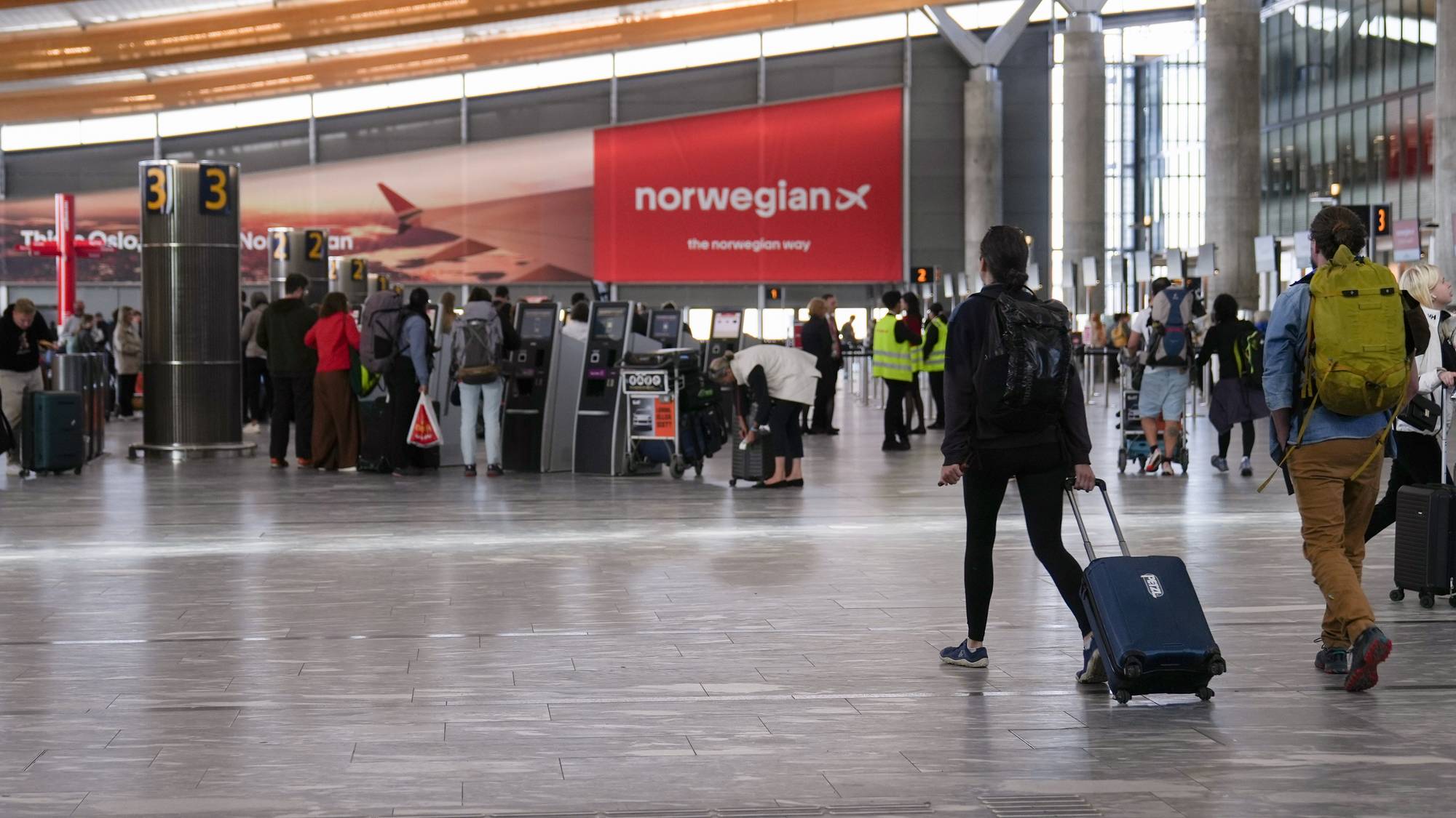 Reisende med bagasje går gjennom en flyplasshall med Norwegian-skilt i bakgrunnen. (Bildebeskrivelsen er laget av en KI-tjeneste)