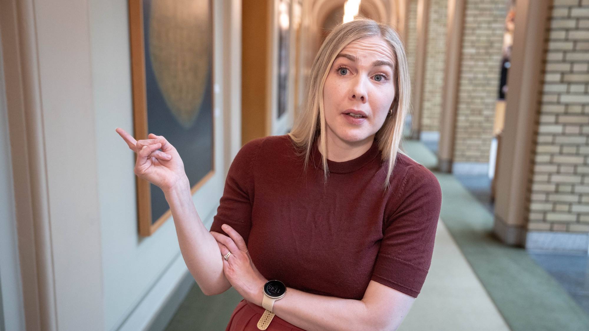 En kvinne med blondt hår peker med pekefingeren mot venstre. Hun har på seg en mørk rød genser og et rødt skjørt. Kvinnen har et ur på venstre håndledd. I bakgrunnen er det en lang korridor med lyse vegger og mursteinsdetaljer i taket. 