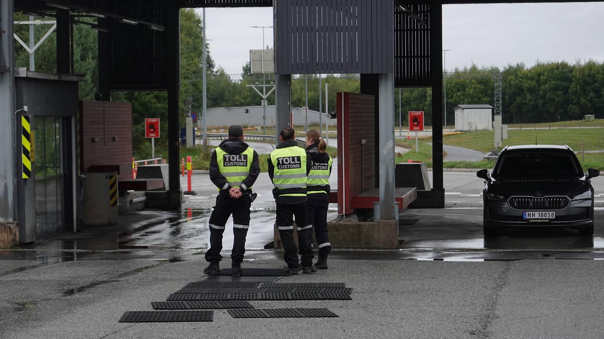Tollbetjenter i refleksvester står ved en tollstasjon med en bil parkert i nærheten. (Bildebeskrivelsen er laget av en KI-tjeneste)