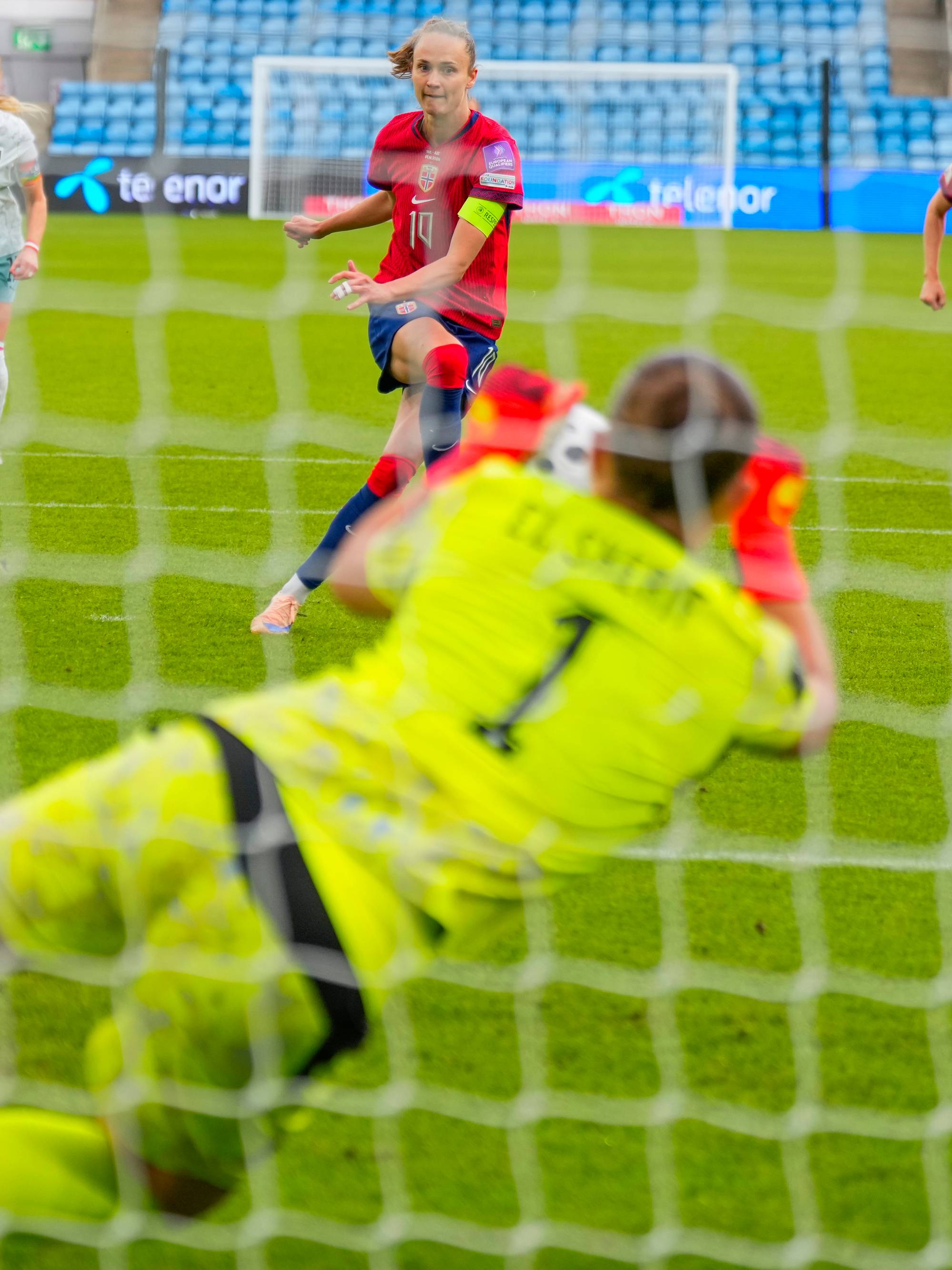 En fotballkeeper i limegrønn drakt dykker mot høyre i et forsøk på å redde et skudd. To fotballspillere i røde drakter er synlige i bakgrunnen. Bildet er tatt fra bak et fotballnett. (Bildebeskrivelsen er laget av en KI-tjeneste)