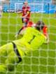Heiko Junge / NTB En fotballkeeper i limegrønn drakt dykker mot høyre i et forsøk på å redde et skudd. To fotballspillere i røde drakter er synlige i bakgrunnen. Bildet er tatt fra bak et fotballnett. (Bildebeskrivelsen er laget av en KI-tjeneste)