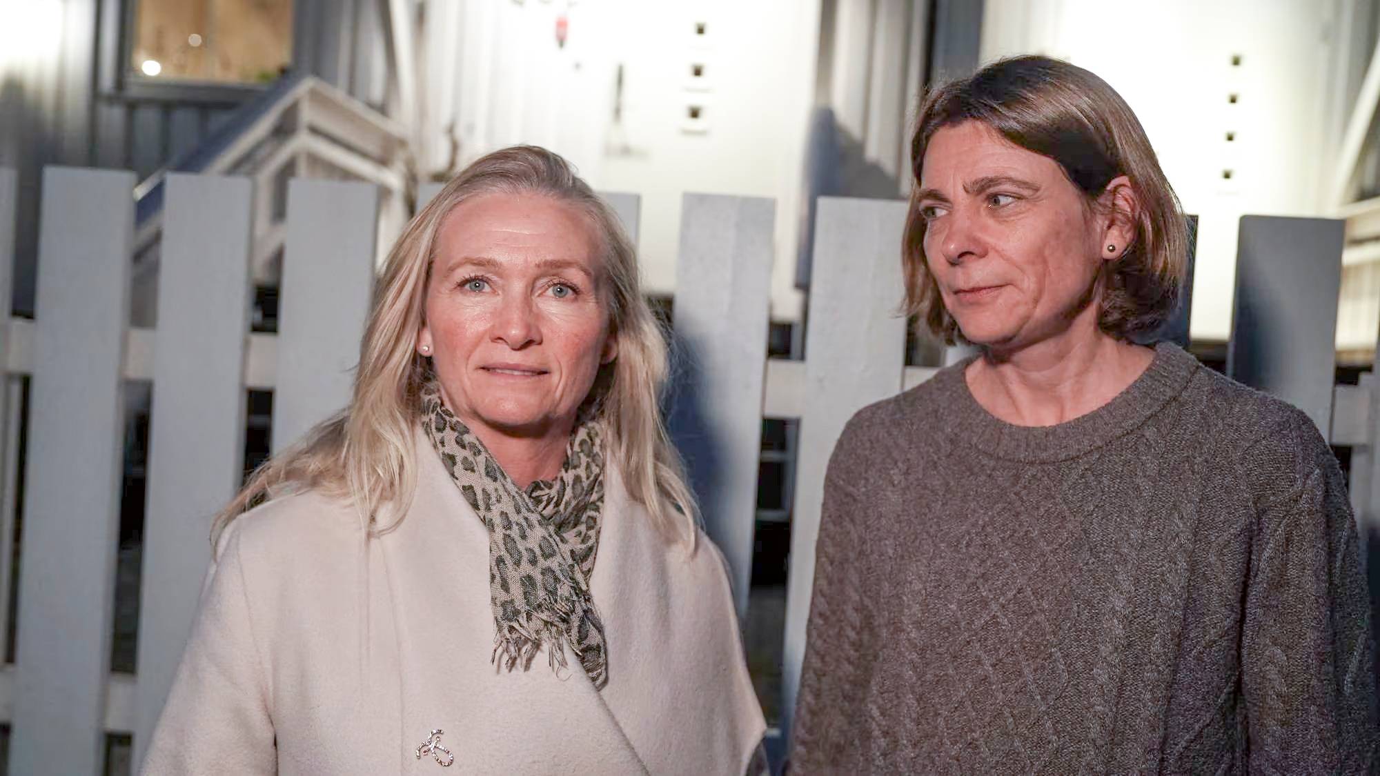 To kvinner står ved siden av hverandre foran et hvitt stakittgjerde. Kvinnen til venstre har blondt hår og en lys frakk med et dyremønstret skjerf. Kvinnen til høyre har brunt hår og en mørkegrå genser. Bak dem er det en bygning med vinduer.