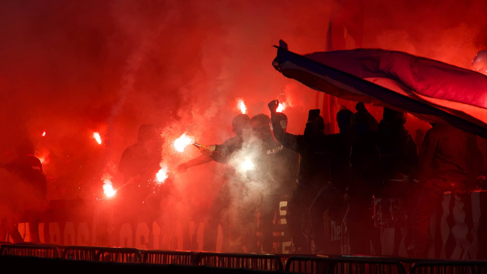 Supportere på en fotballtribune tenner bluss, noe som skaper kraftig rød røyk og lys. (Bildebeskrivelsen er laget av en KI-tjeneste)