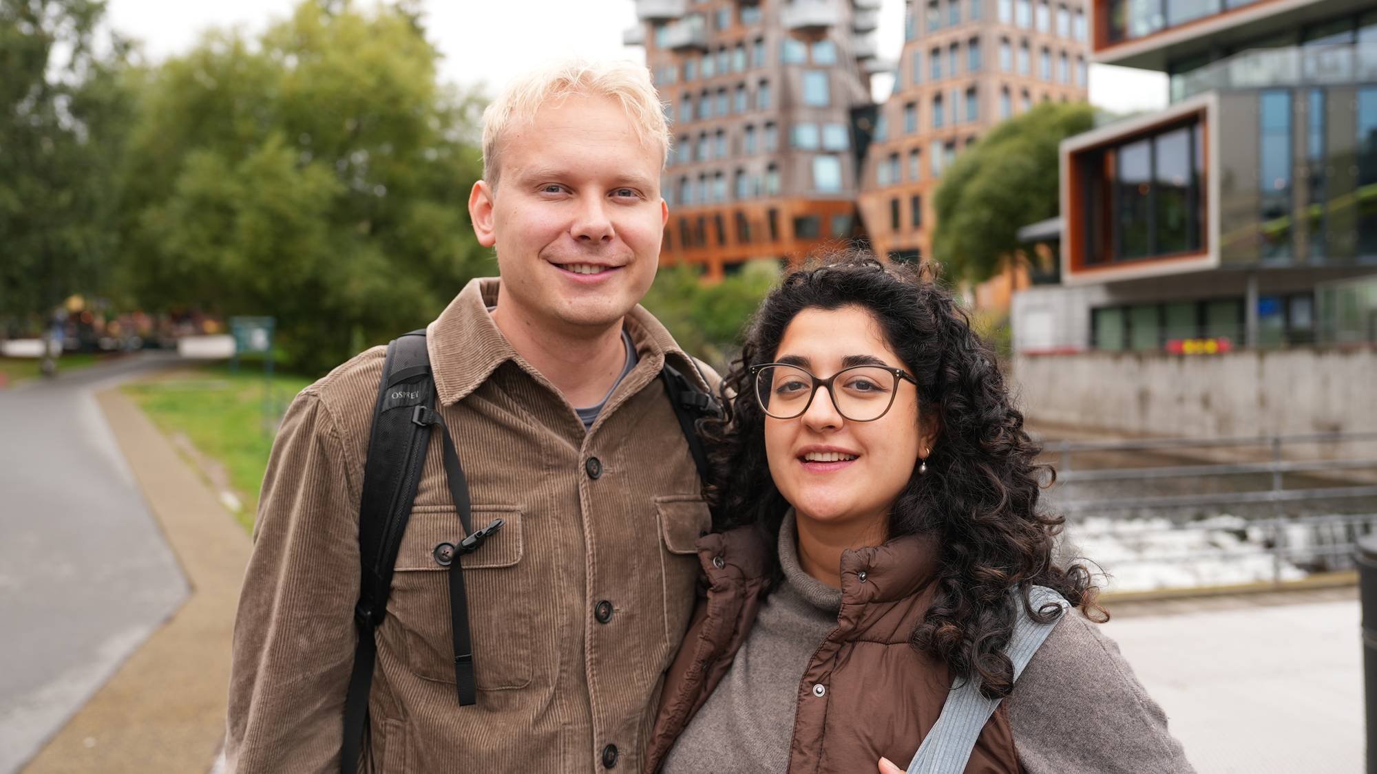 En lyshåret mann og en kvinne med mørkt krøllet hår står sammen utendørs foran moderne bygg. (Bildebeskrivelsen er laget av en KI-tjeneste)