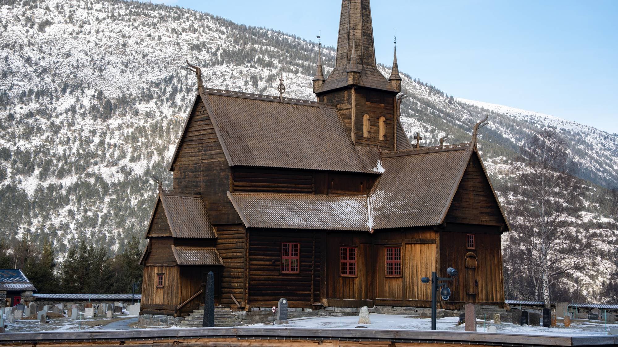Bildet viser en stavkirke med karakteristisk tretak og spiss tårn. Kirken er plassert i et vinterlandskap, omgitt av snødekte fjell. I forgrunnen er det en gravlund med flere gravsteiner synlige. Himlingen er klar med få skyer, noe som gir et rolig uttrykk. (Bildebeskrivelsen er laget av en KI-tjeneste)