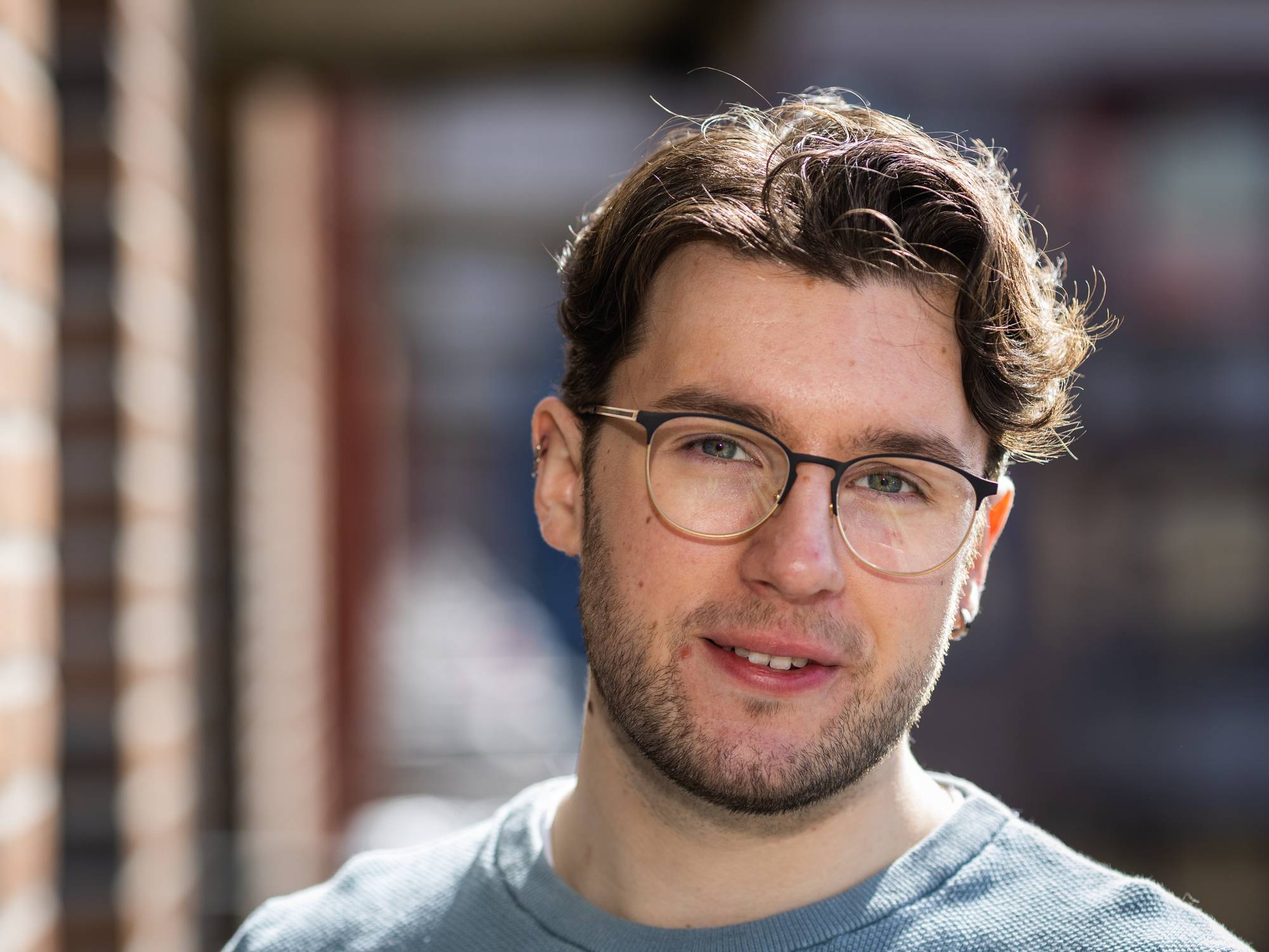 Et annet portrett av Daniel Restad. Han står på balkongen med solen i nakken, og ser undrende i kamera mens han smiler subtilt.
