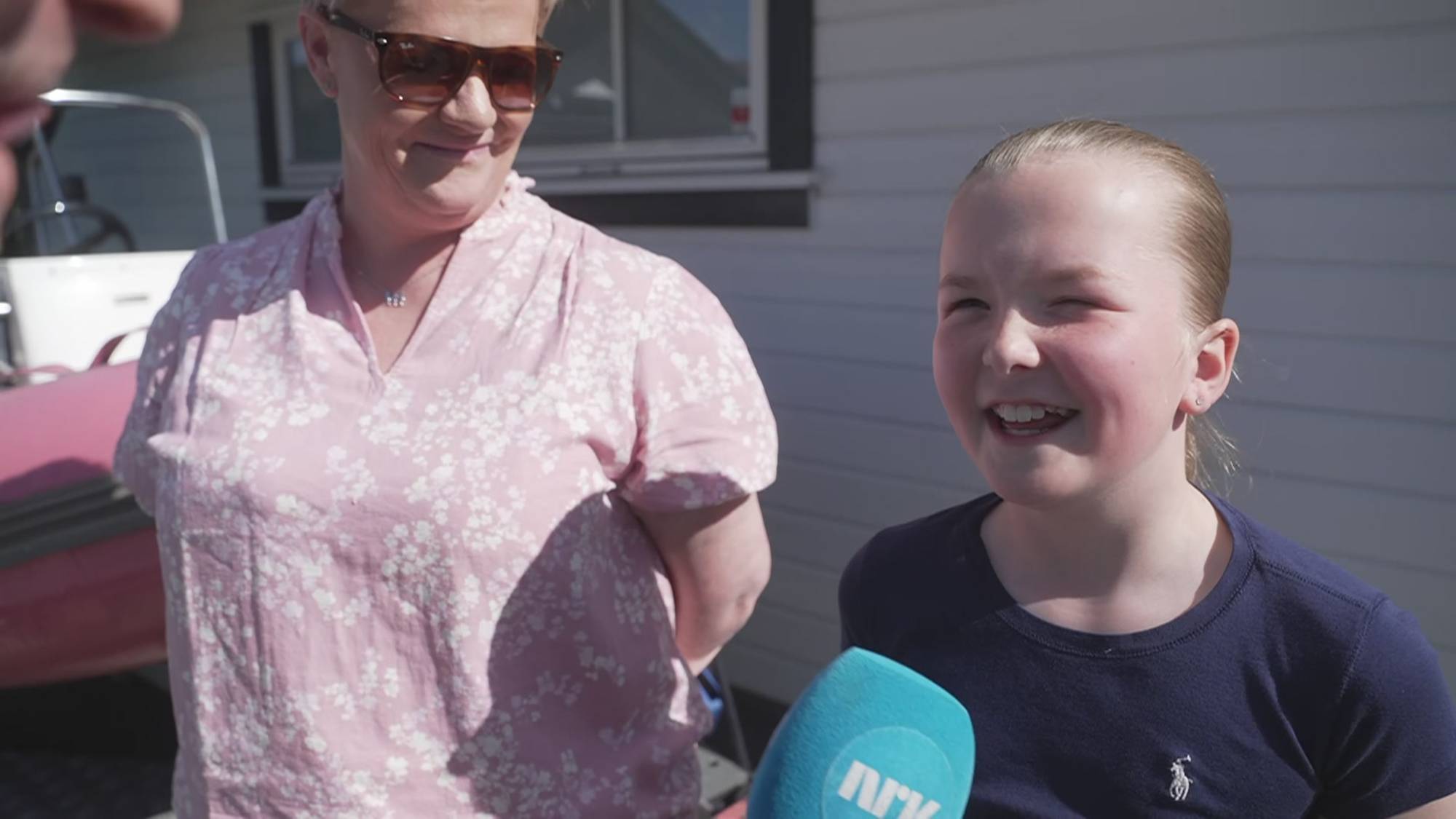 En ung jente blir intervjuet. Hun står foran en kvinne i en rosa blomstrete bluse. Begge er ute i solskinn, med en bygning i bakgrunnen. En mikrofon med NRK-logo er synlig. (Bildebeskrivelsen er laget av en KI-tjeneste)