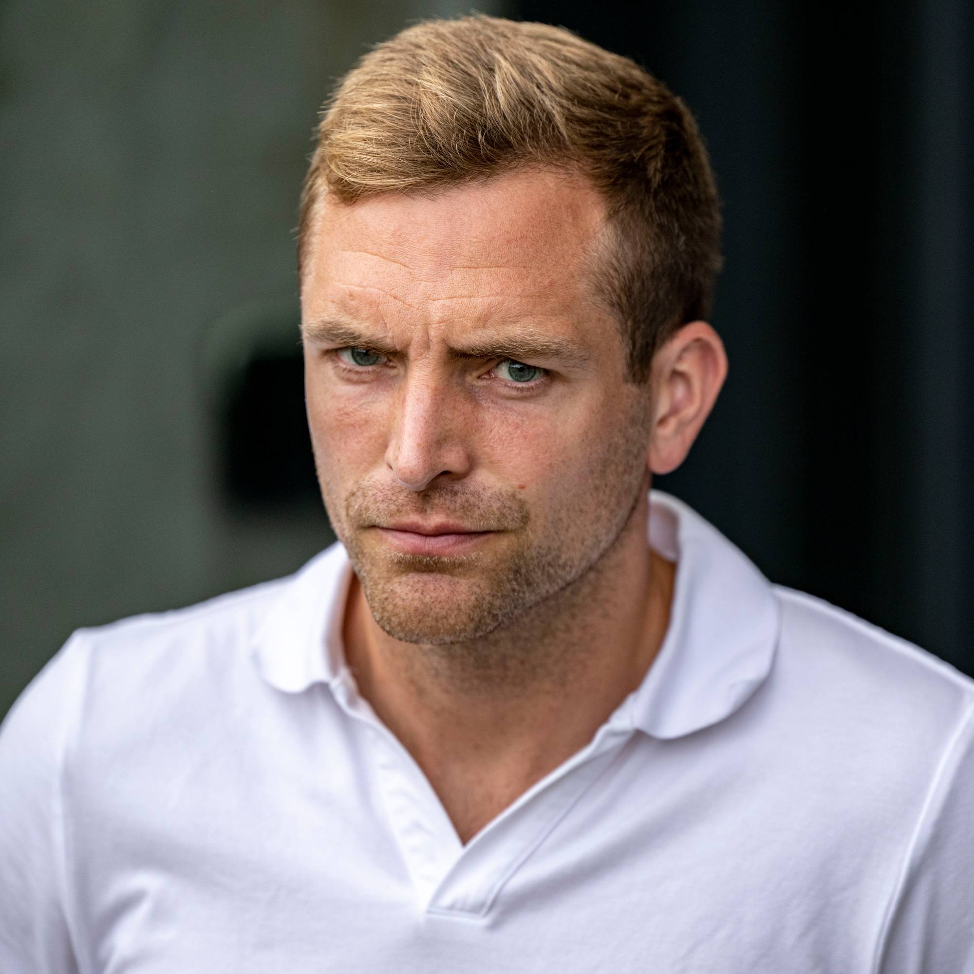 Mann med kort blondt hår og skjegg i en hvit pikétrøye. Han ser mot venstre. Bakgrunnen er mørk. (Bildebeskrivelsen er laget av en KI-tjeneste)