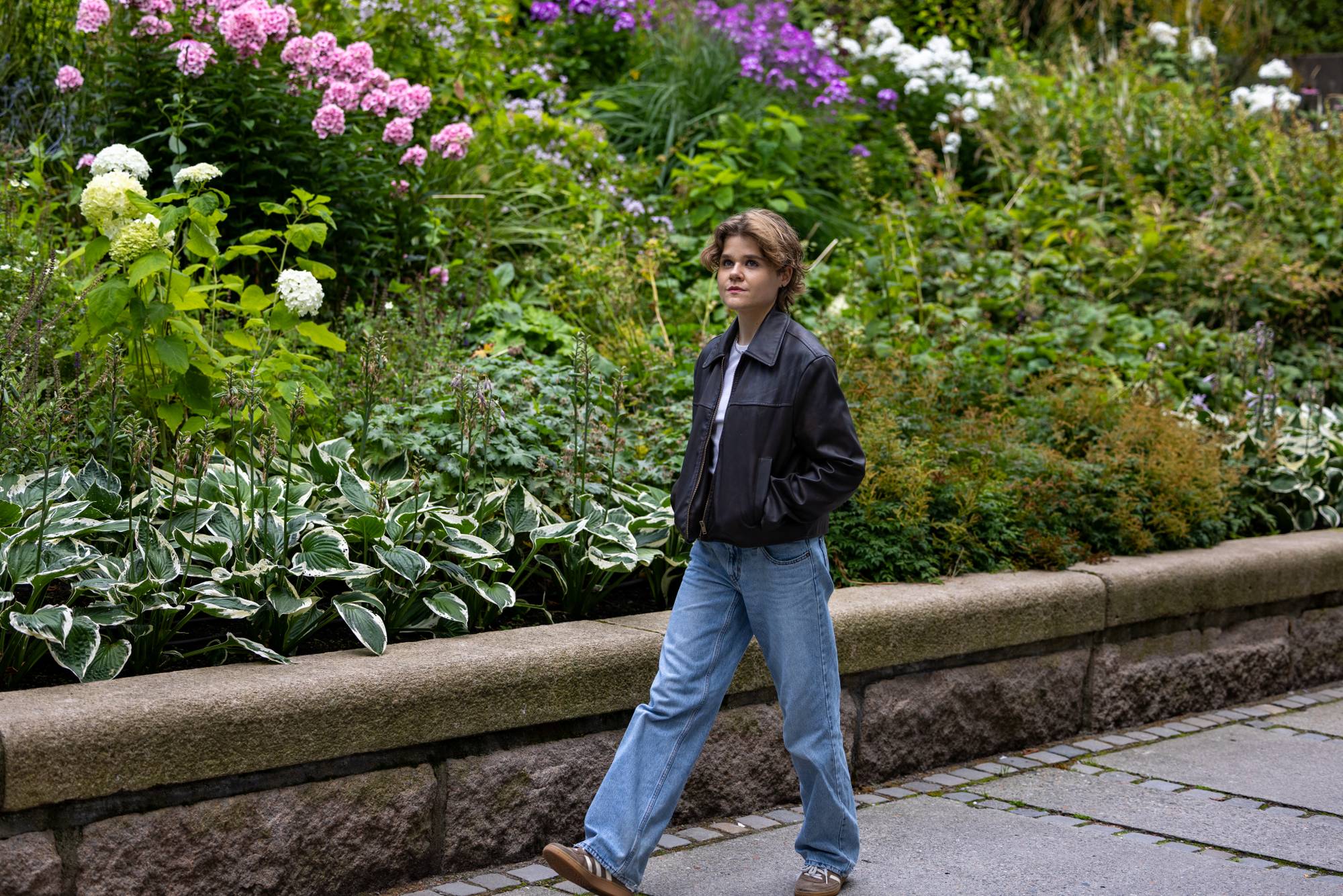 Ung kvinne med svart skinnjakke går forbi en blomsterhage i en park.