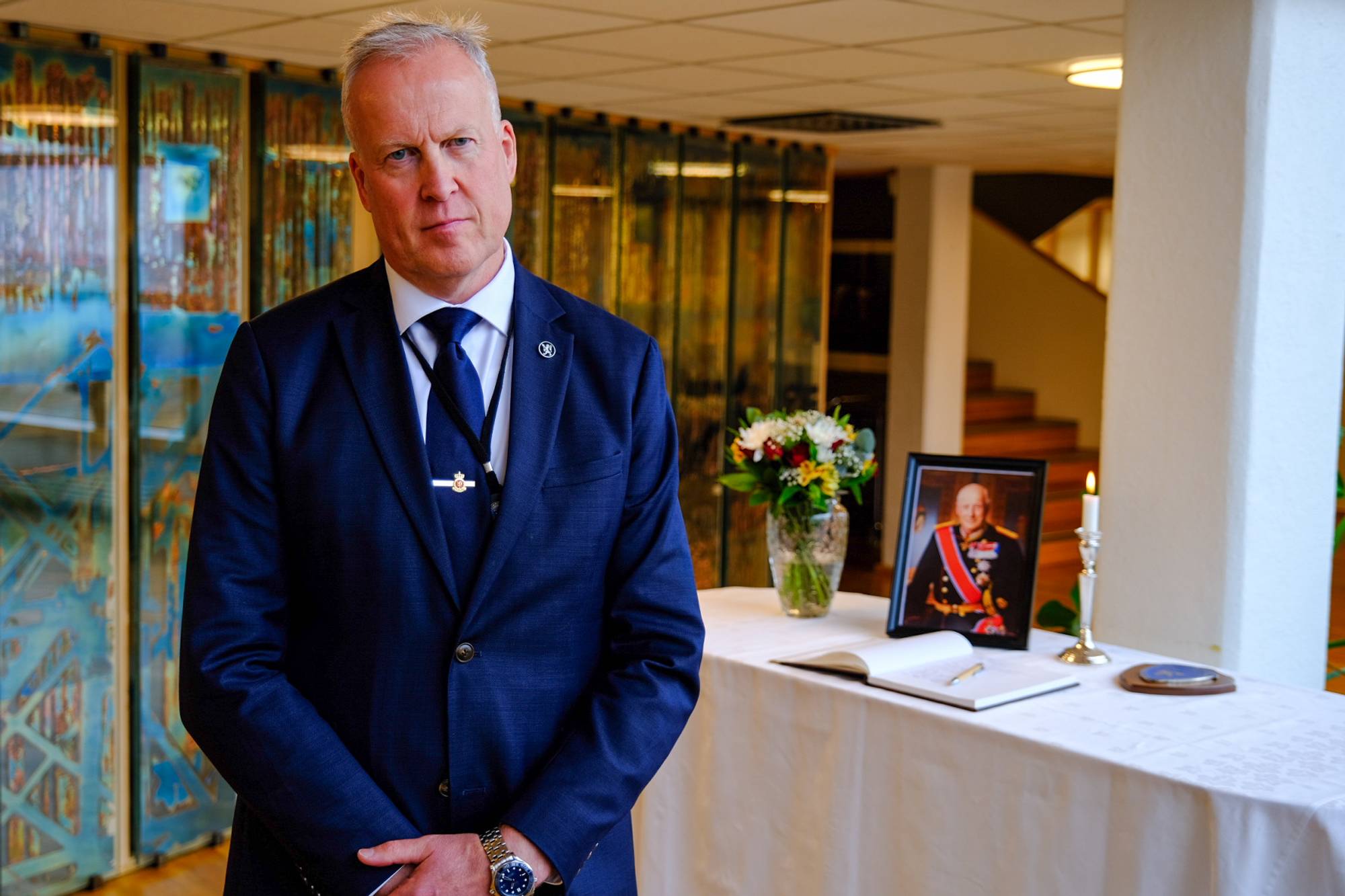 Mann i dress står foran et bord med et portrett av kong Harald, en kondolanseprotokoll og et lys. (Bildebeskrivelsen er laget av en KI-tjeneste)