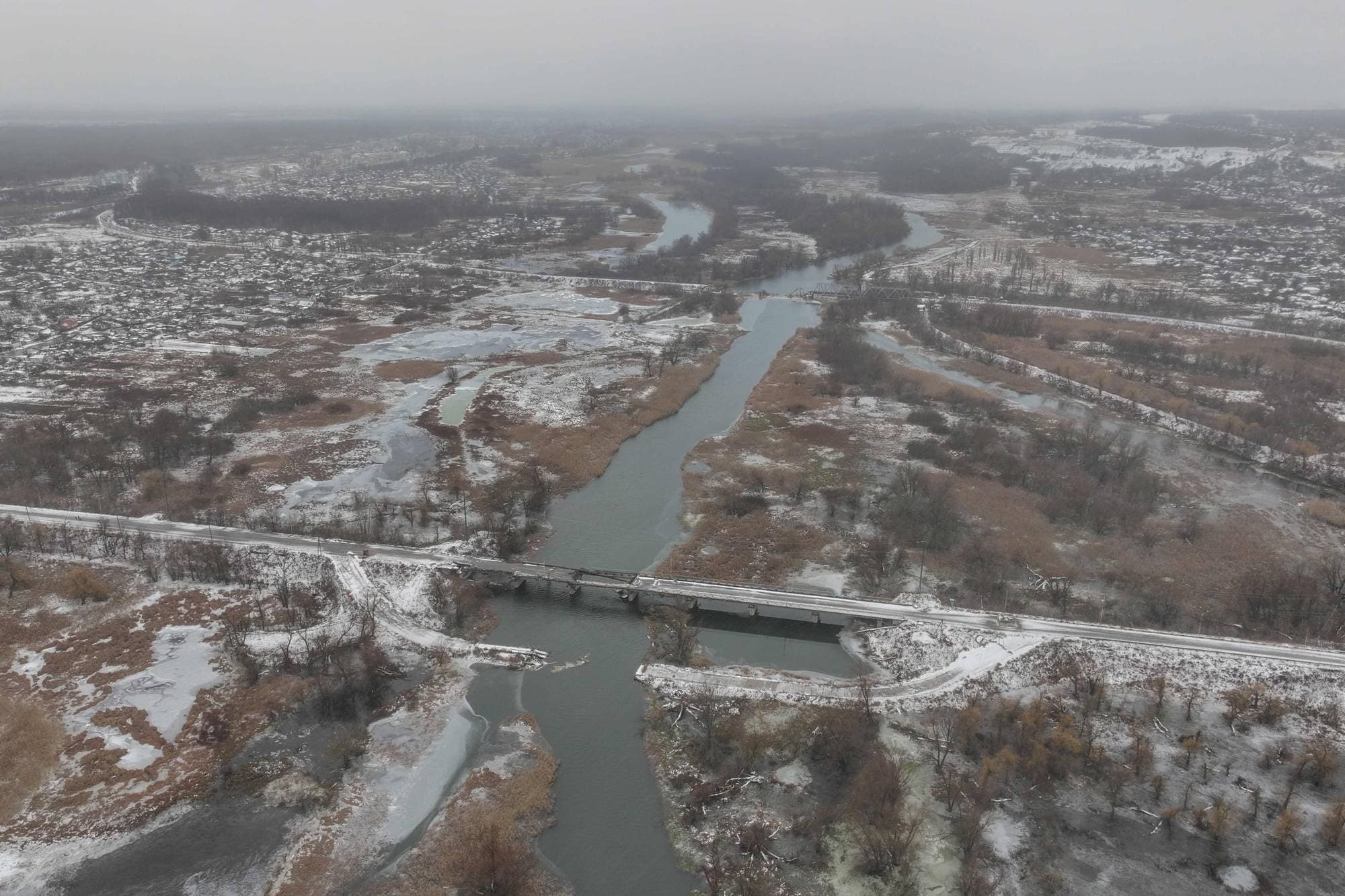 Dronebilde viser en elv som renner gjennom et snødekt landskap. En bro krysser elven, med en vei og jernbanespor som strekker seg langs bredden. I bakgrunnen ligger et tettsted og et skogkledd åslandskap under en grå himmel. (Bildebeskrivelsen er laget av en KI-tjeneste)