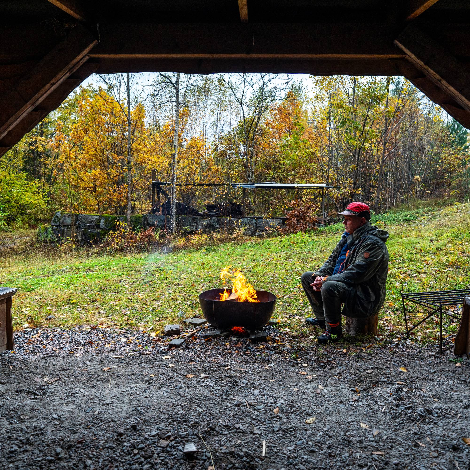 En mann sitter ved et bål i et trebygg med utsikt mot en høstskog. (Bildebeskrivelsen er laget av en KI-tjeneste)