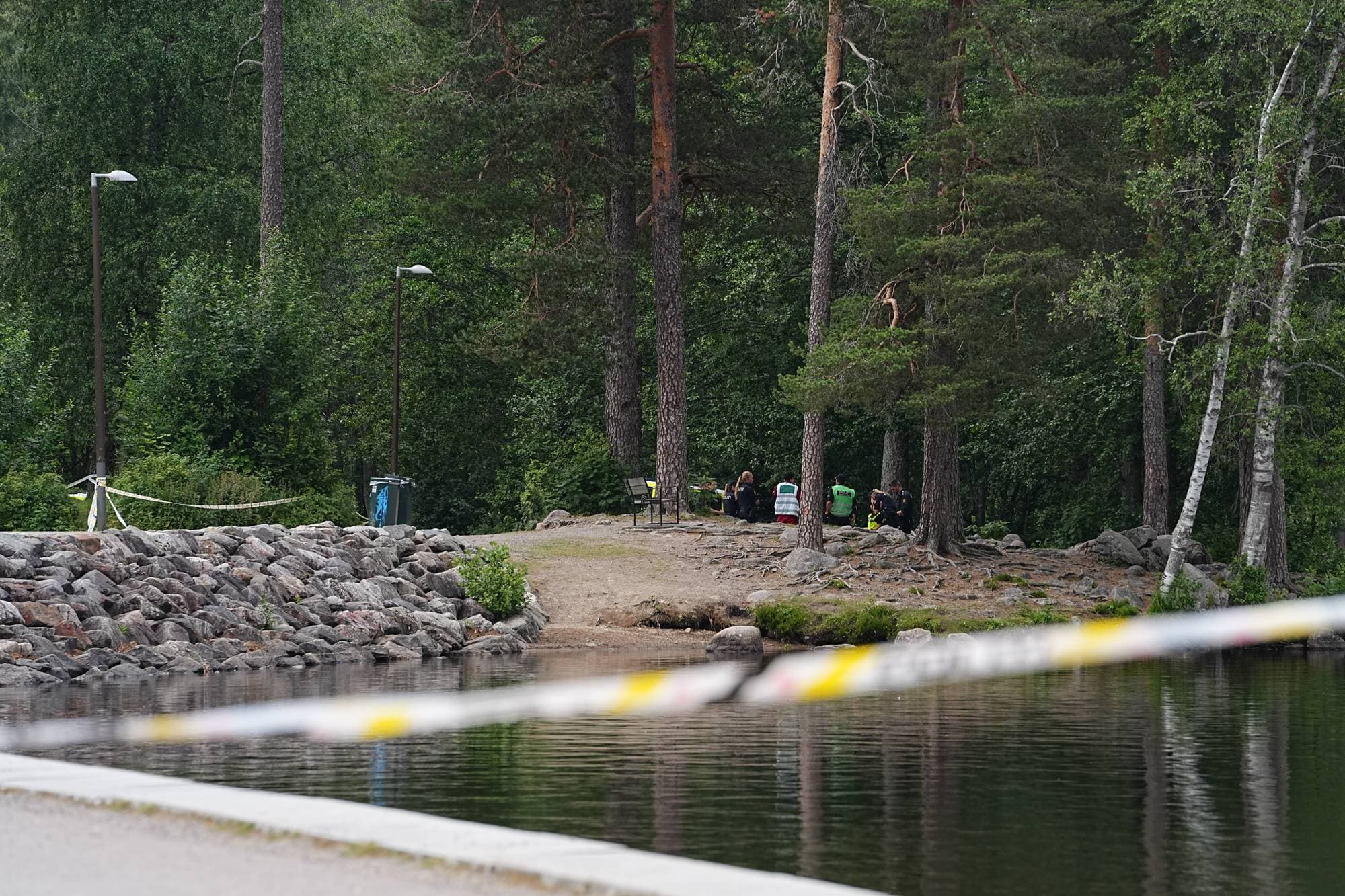 Politi og redningspersonell ved et avsperret skogsområde ved vannkanten. (Bildebeskrivelsen er laget av en KI-tjeneste)