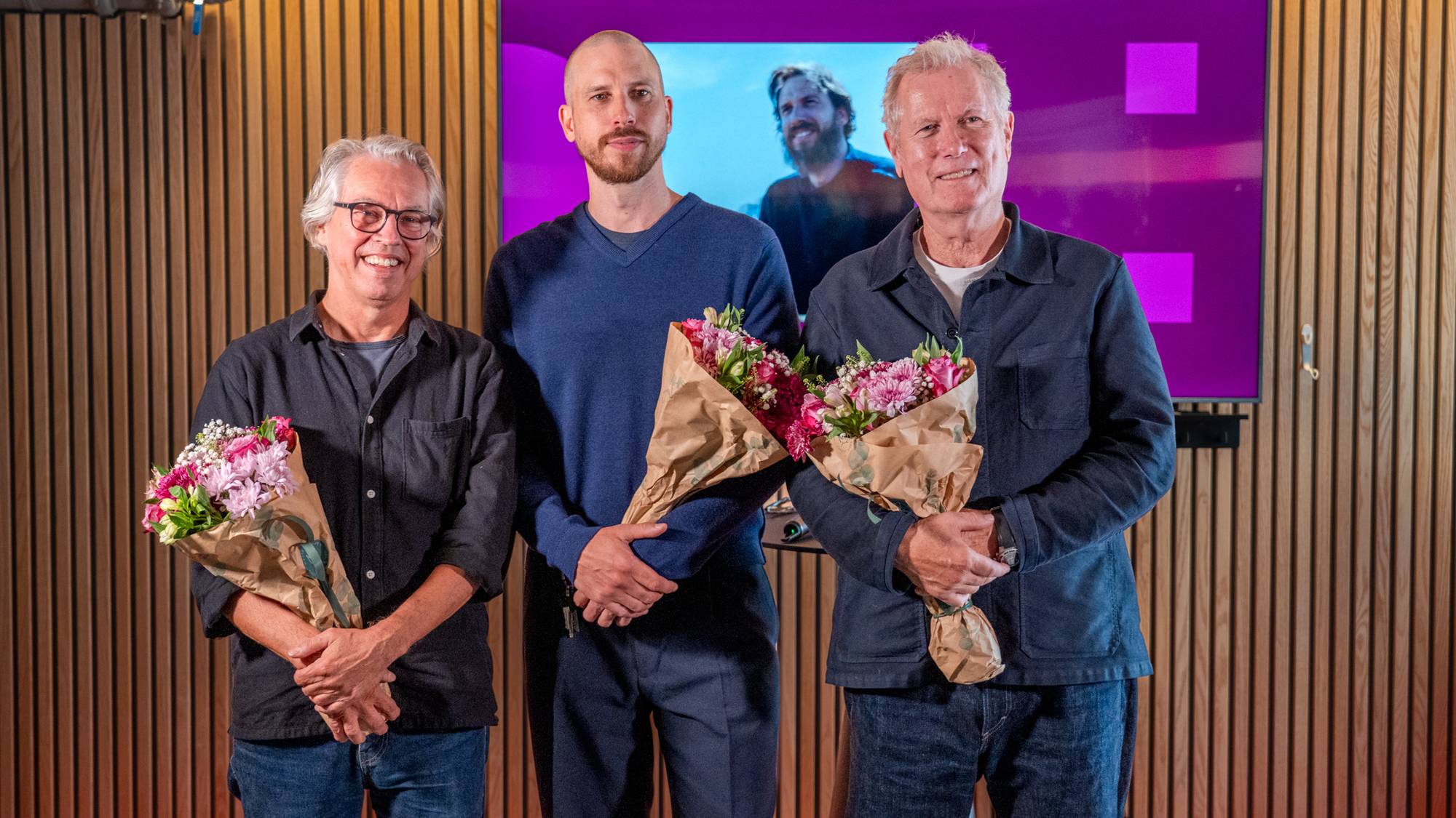 Tre menn som står med blomsterbuketter og ser i kameraet