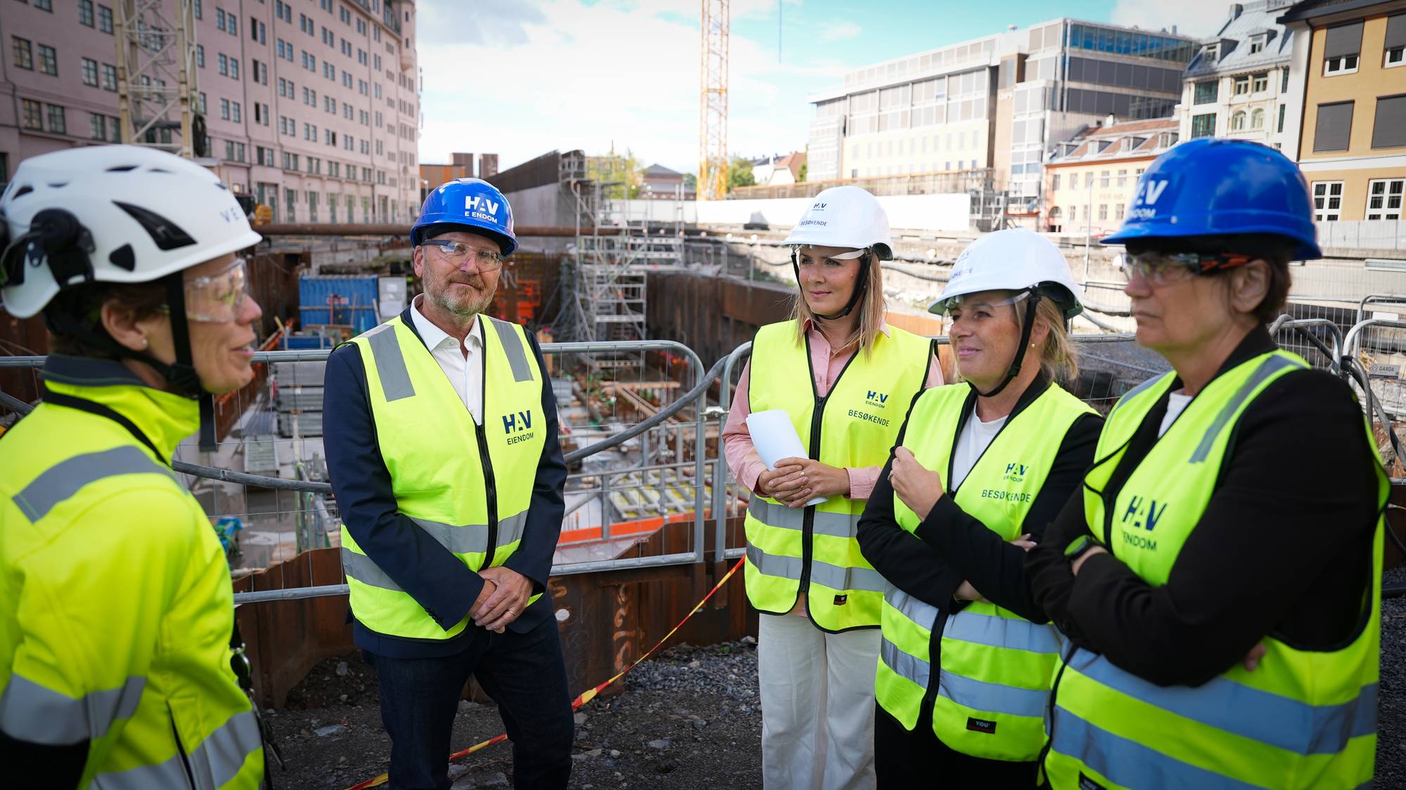 Fem personer i verneutstyr står ved en byggeplass og samtaler. 