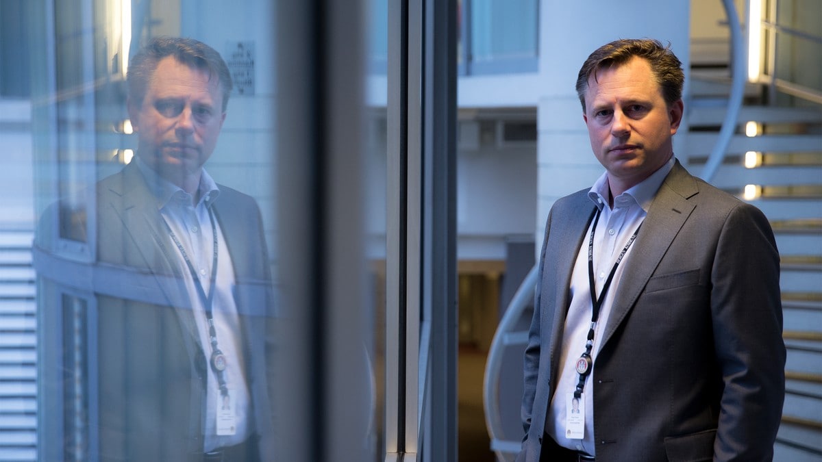 En mann i grå dress står i en moderne bygning. Han har en lanyard med nøkkelkort og ser mot kameraet. Refleksjonen hans kan sees i glasset foran ham. I bakgrunnen er det en spiraltrapp og interiørdetaljer. FOTO: Simen Sundfjord Otterlei