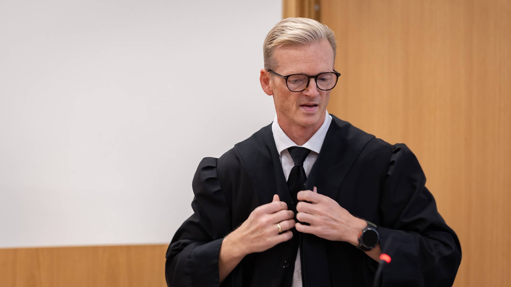 En mann iført svart kappe og slips er i en rettssal. Mannen har briller med mørk innfatning. Han retter på kappen