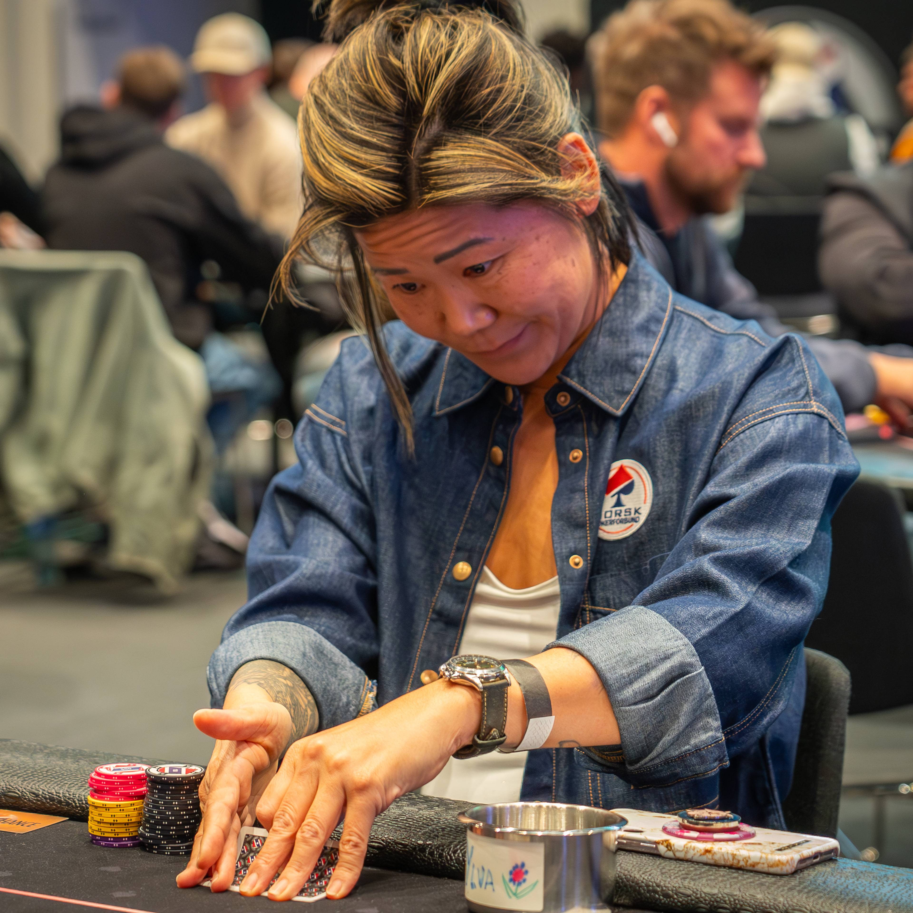 Kvinne i olieskjorte spiller poker ved et bord med sjetonger i et travelt rom.