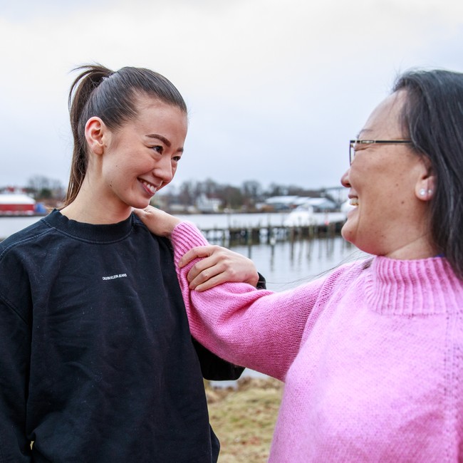 Datter Emilie Lervik iført svart treningstøy, og mor Regine Kim Lervik iført rosa ullgenser og briller, holder hverandres håns og ser på hverandre smilende foran Glomma i Lisleby.