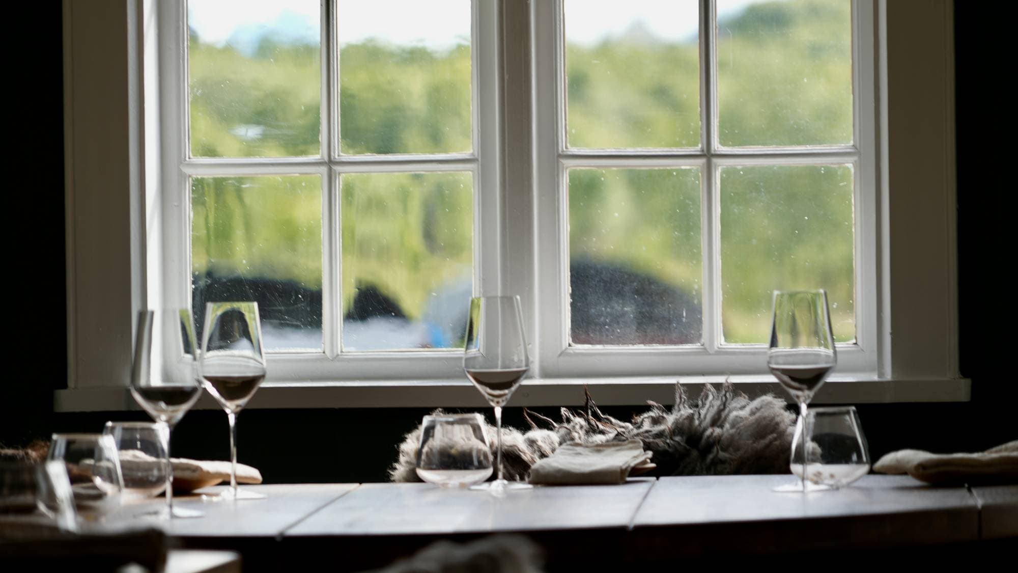 Seks vinglass står på et bord foran et vindu. Utenfor vinduet er det grønt landskap. Bordet er dekket med en duk og noen ulltekstiler. (Bildebeskrivelsen er laget av en KI-tjeneste)