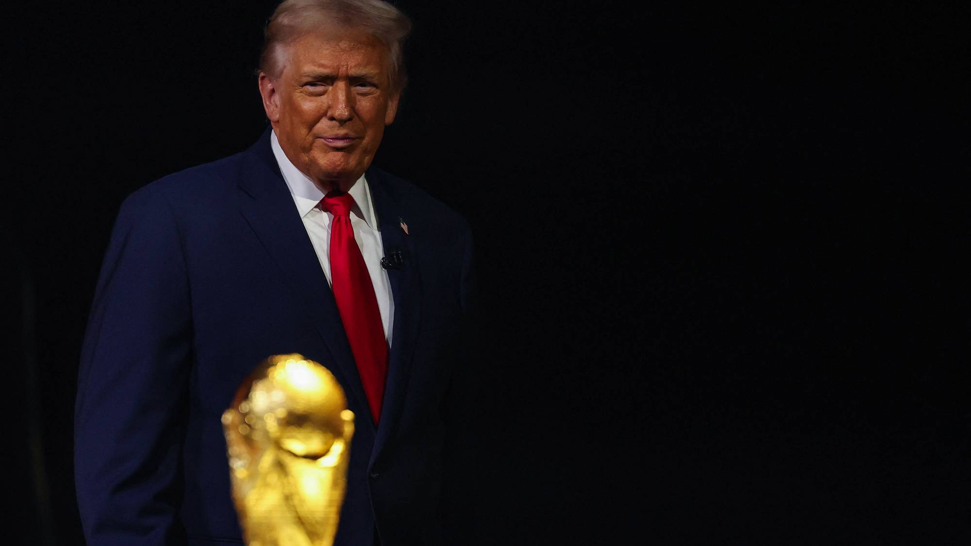 Donald Trump i mørk dress og rødt slips mot en sort bakgrunn. Et uklart gulltrofé står i forgrunnen. (Bildebeskrivelsen er laget av en KI-tjeneste)
