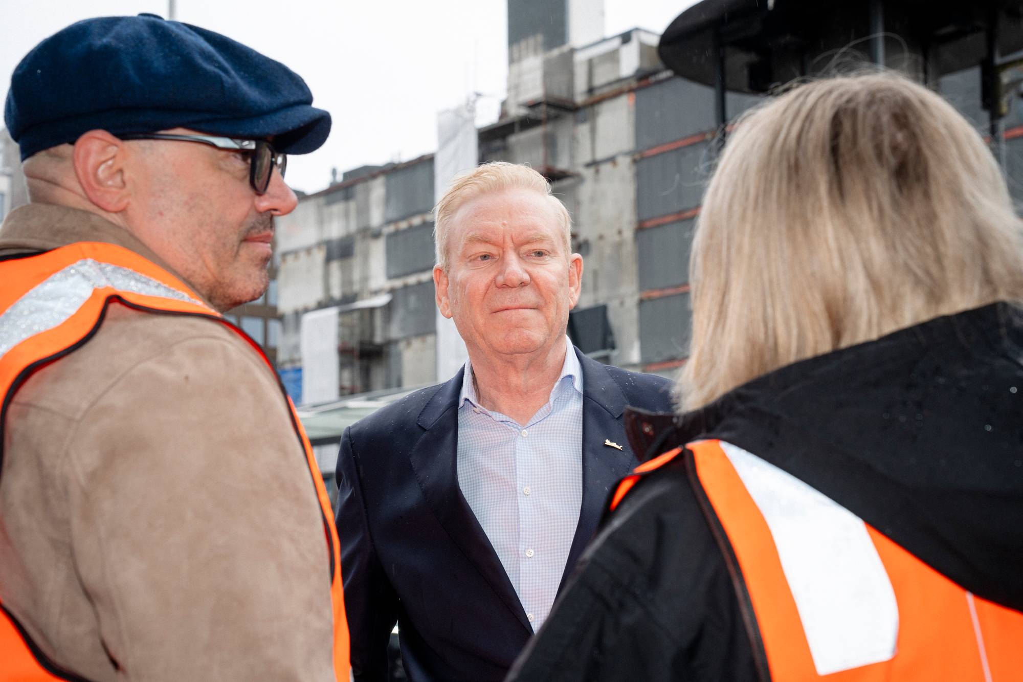 En mann i dress snakker med to personer ikledd refleksvester. Mannen i dress har et lyseblått rutete skjorte. Bak dem sees en bygningsfasade med mange bokser. (Bildebeskrivelsen er laget av en KI-tjeneste)
