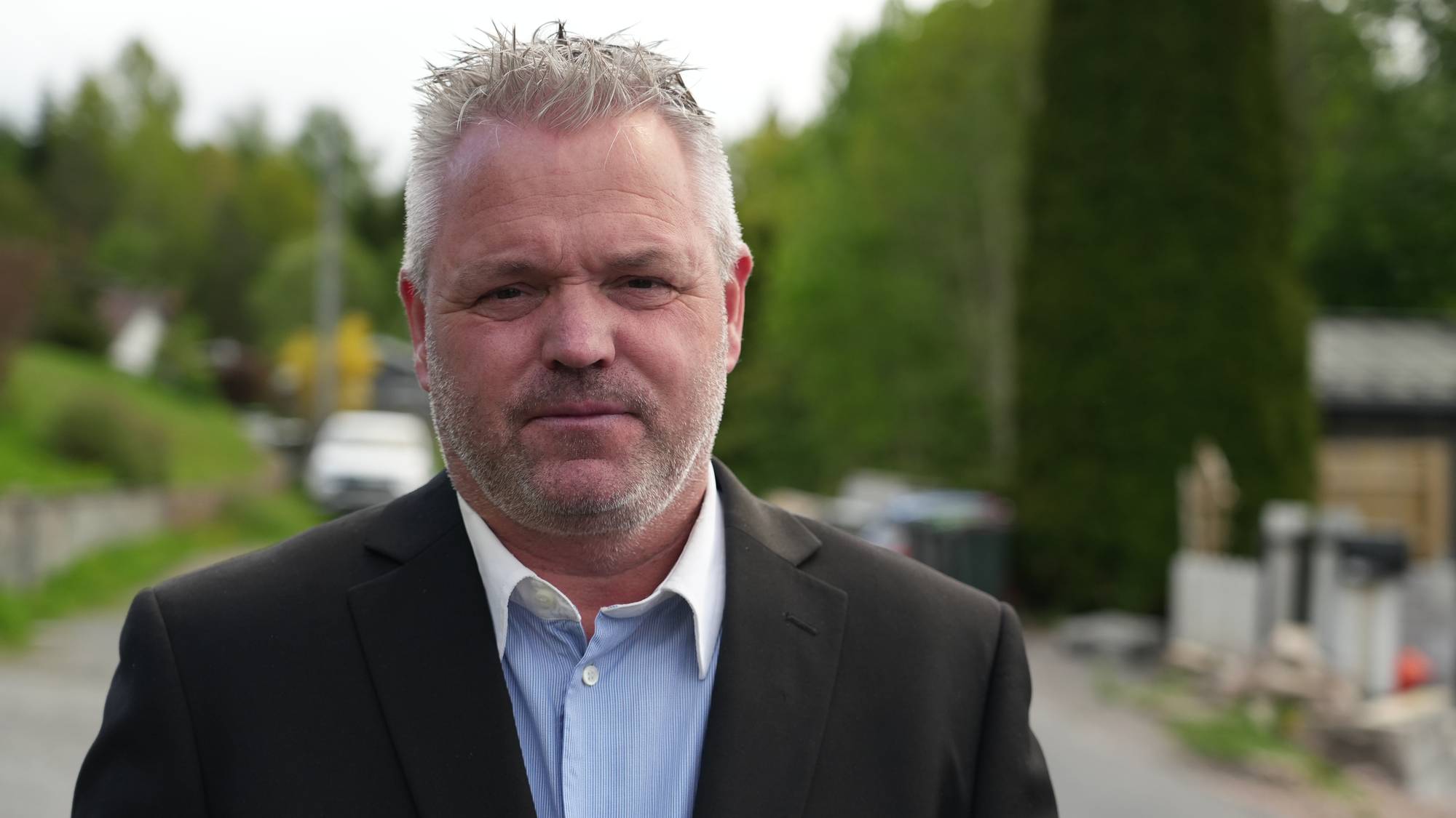 En mann i svart dressjakke og blå skjorte ser mot kamera. Bak ham er det grønt løvverk og en vei.
