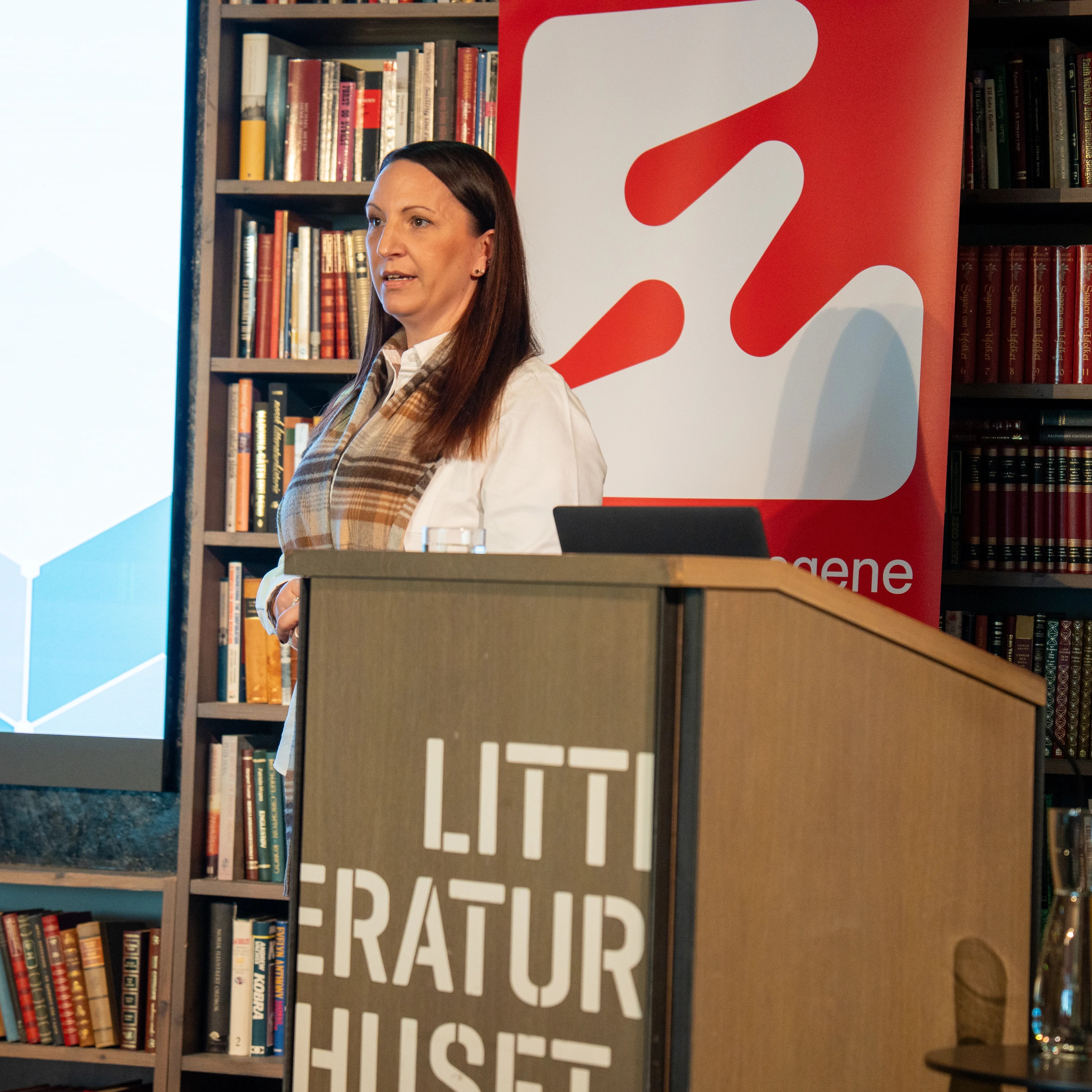 Kvinne holder foredrag fra en talerstol i et rom med bokhyller. En prosjektør viser en presentasjon. (Bildebeskrivelsen er laget av en KI-tjeneste)