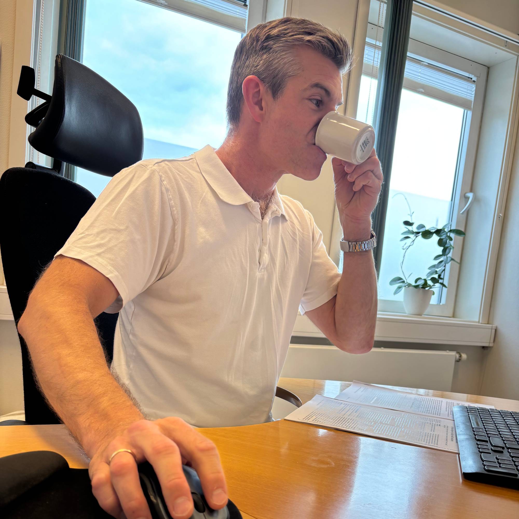 Mann drikker kaffe mens han holder hånden på musepekeren på dataen.