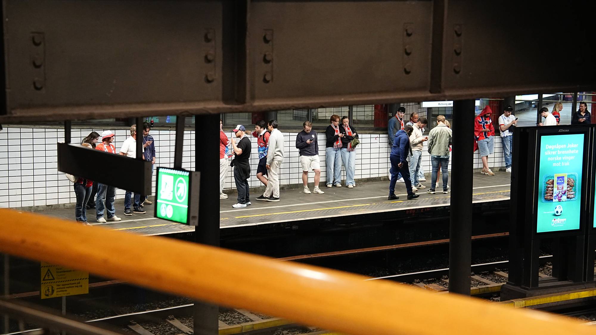 Folk står på en perrong på en t-banestasjon. Flere av personene har på seg supporterutstyr for et idrettslag. Noen ser på telefonen mens de venter. En stor reklameskjerm med et fotballmotiv vises på høyre side av bildet.