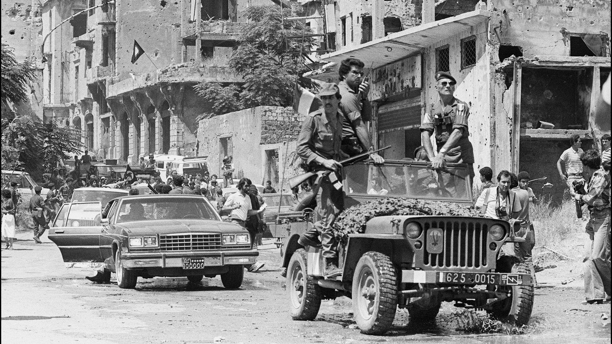 Menn i militæruniform sitter i en militærjeep. En av mennene står oppreist med et gevær. I bakgrunnen er ødelagte bygninger. Bilen har et kamerasymbol på panseret. Det er flere sivile til stede. (Bildebeskrivelsen er laget av en KI-tjeneste)