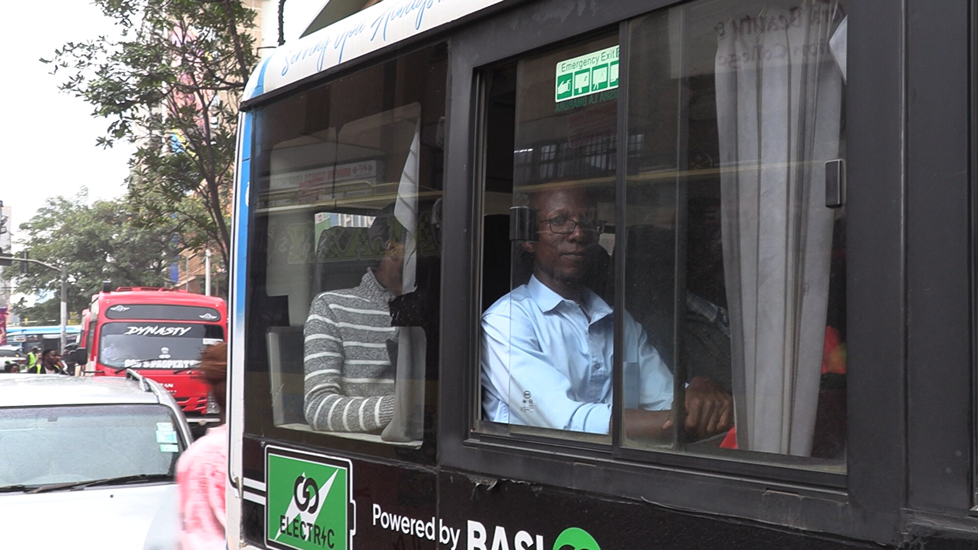 Ein afrikansk mann sit inni ein buss og kikkar mot kamera gjennom vindauget.