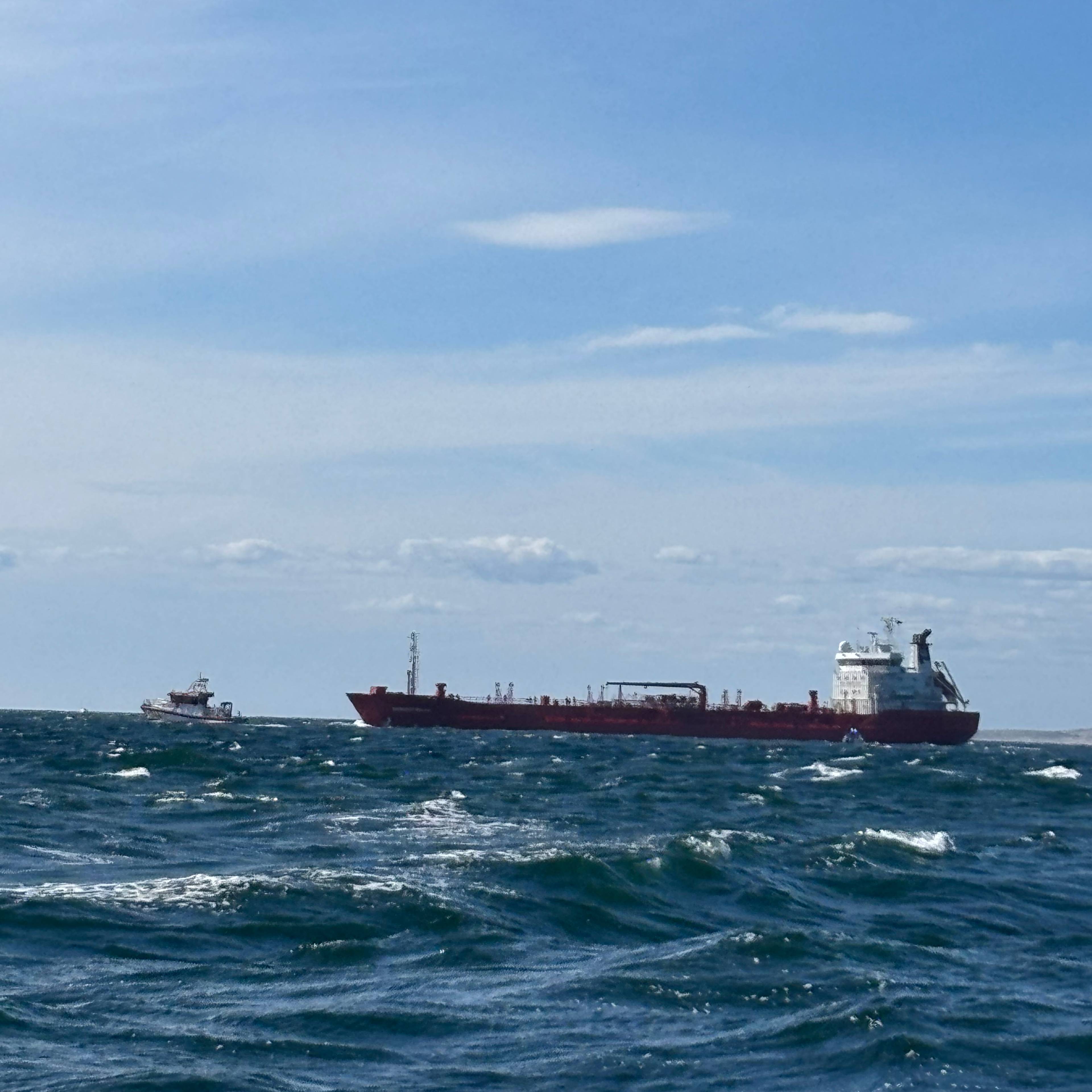 Et rødt tankskip seiler på åpent hav med en mindre båt i nærheten under en blå himmel.
