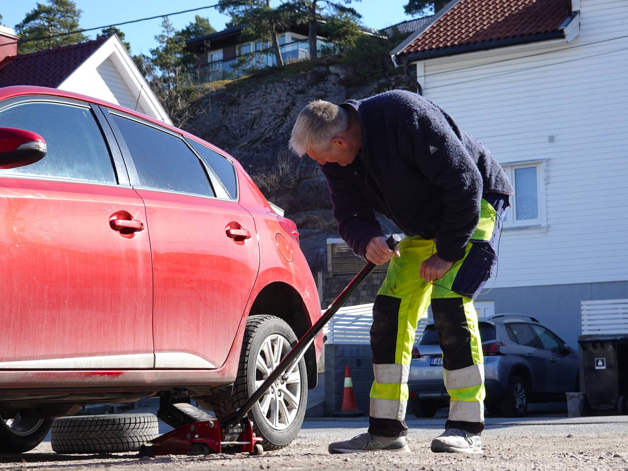 En mann bytter dekk på en rød bil med jekk. Mannen har på seg reflekterende bukser. En reservehjul ligger under bilen. I bakgrunnen står et hvitt hus og flere andre biler. (Bildebeskrivelsen er laget av en KI-tjeneste)