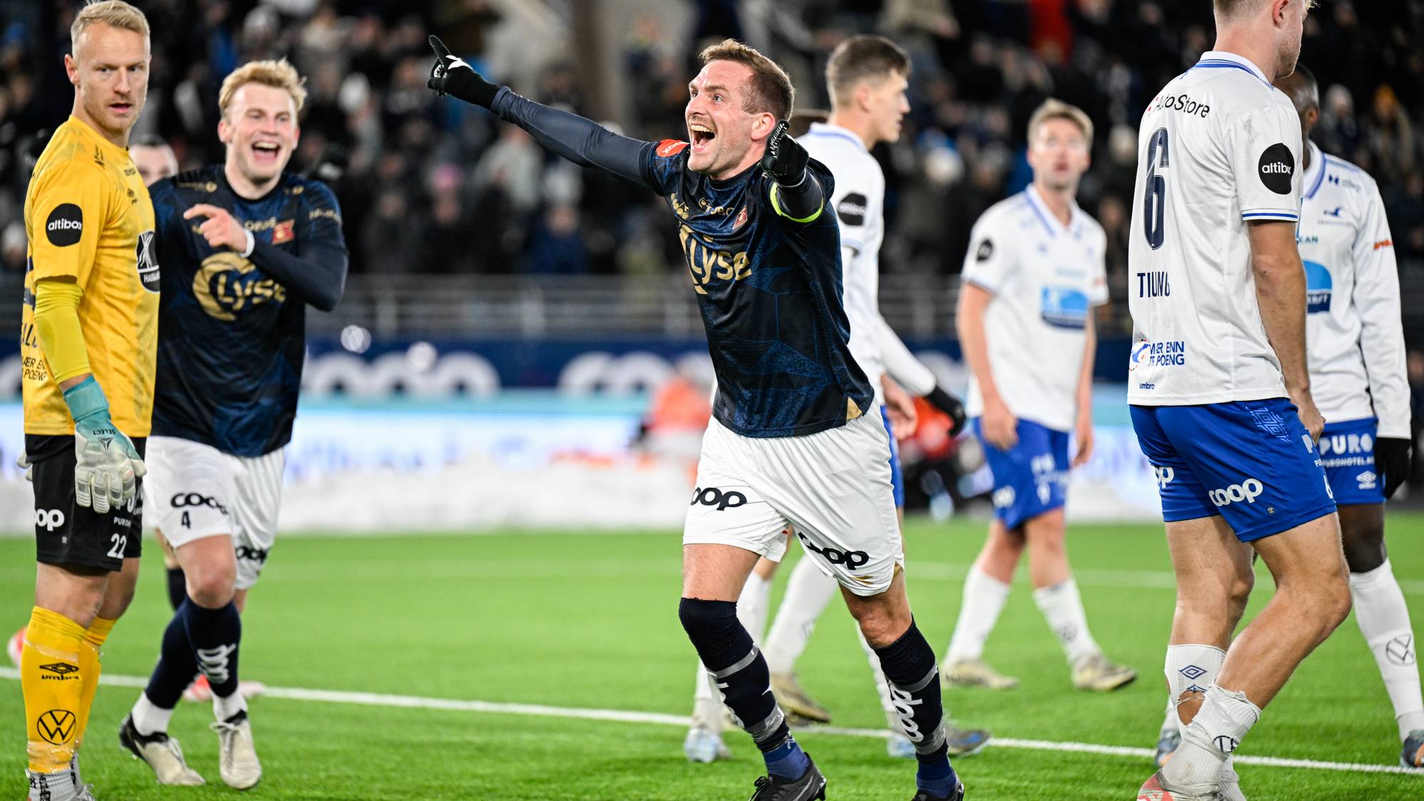 En fotballspiller i mørkeblå drakt jubler med armene hevet på en gressbane. Andre spillere i henholdsvis mørkeblå og hvit drakt ser på. En keeper i gul drakt står til venstre. (Bildebeskrivelsen er laget av en KI-tjeneste)